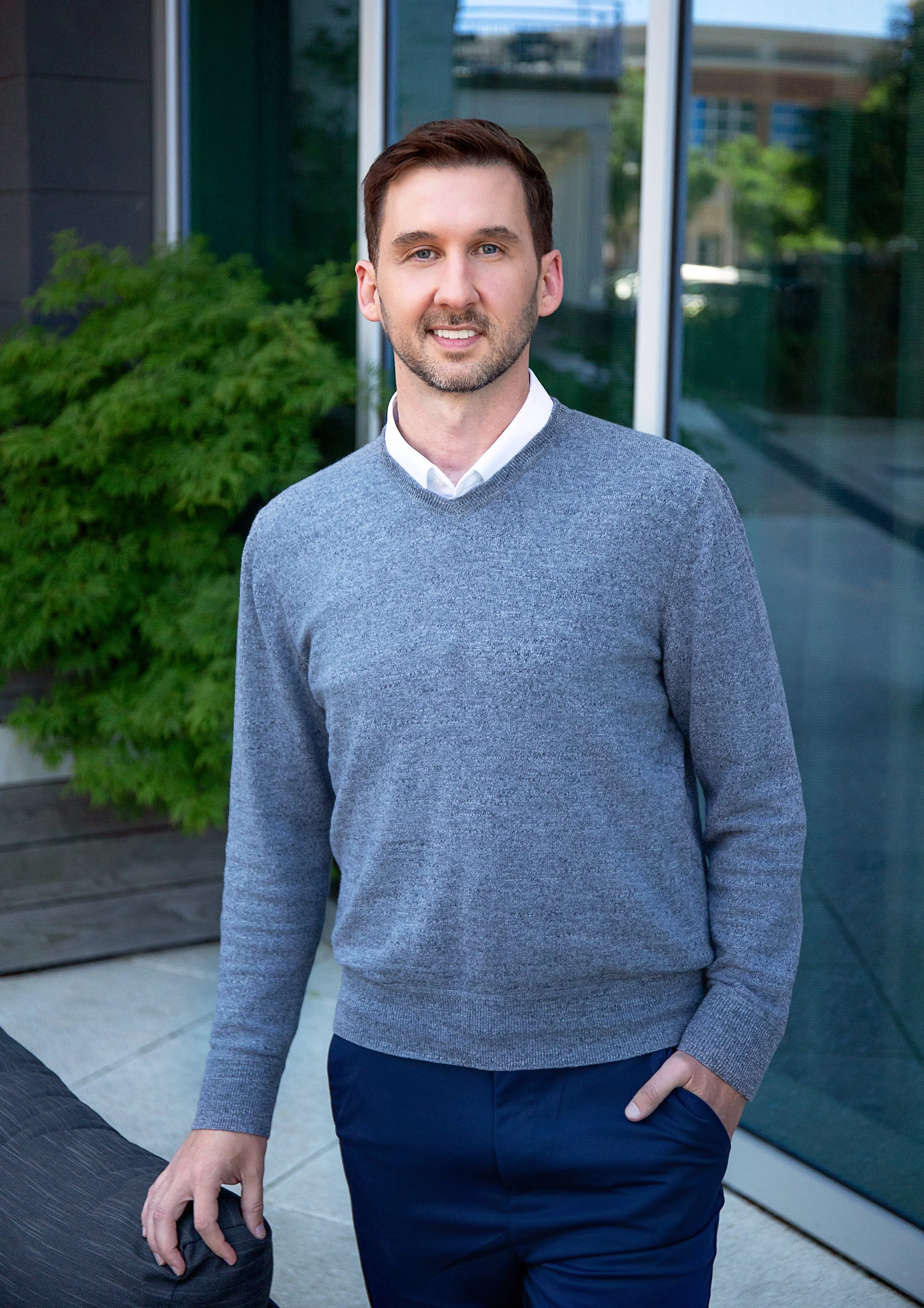 A young man with short dark hair and a beard, wearing a white shirt under a gray sweater, stands outdoors in front of a large glass window with green plants and a building reflected in the background, smiling at the camera.