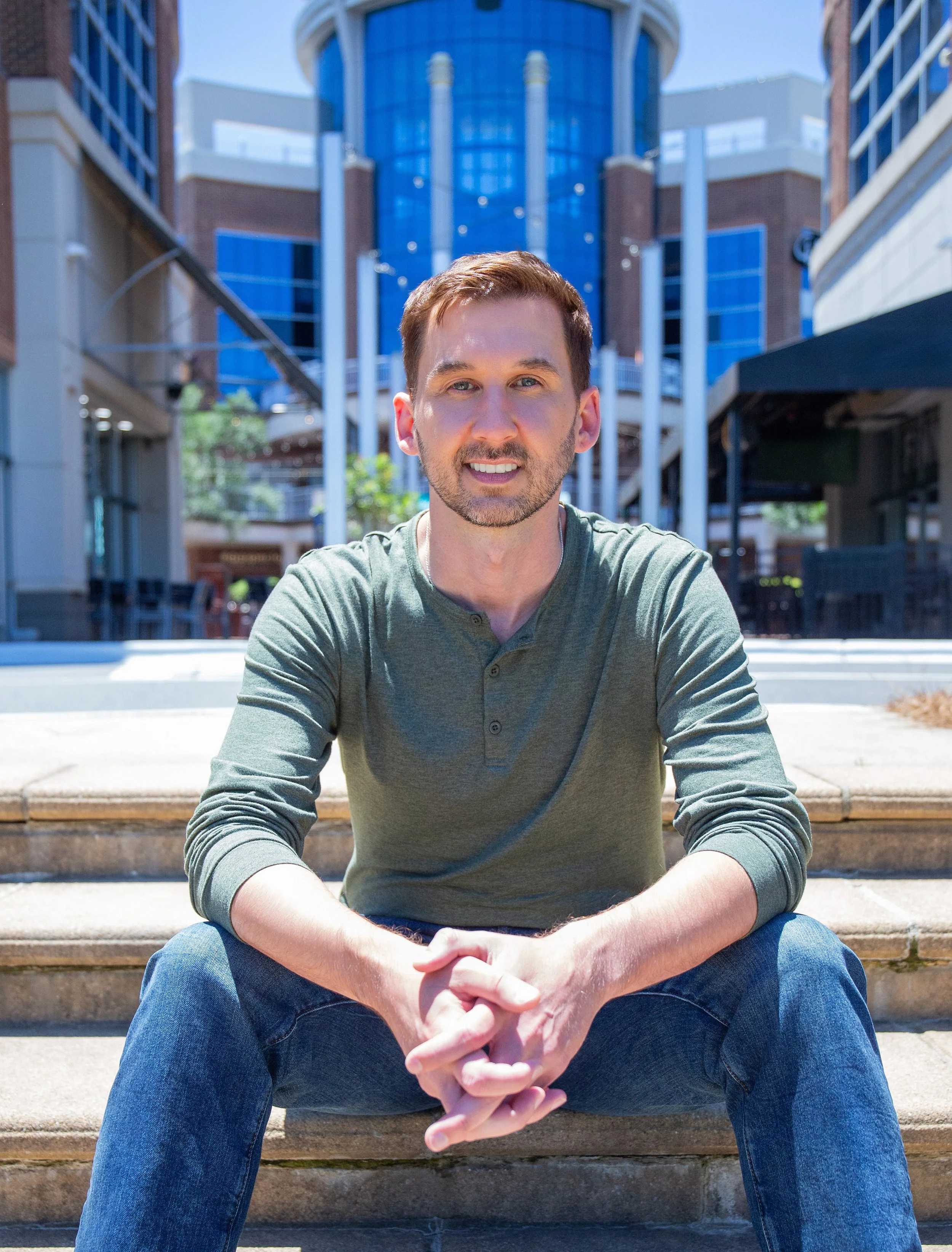 A young man with brown hair and a beard sitting on concrete stairs outdoors, in front of a modern building with glass and brick elements, smiling at the camera.