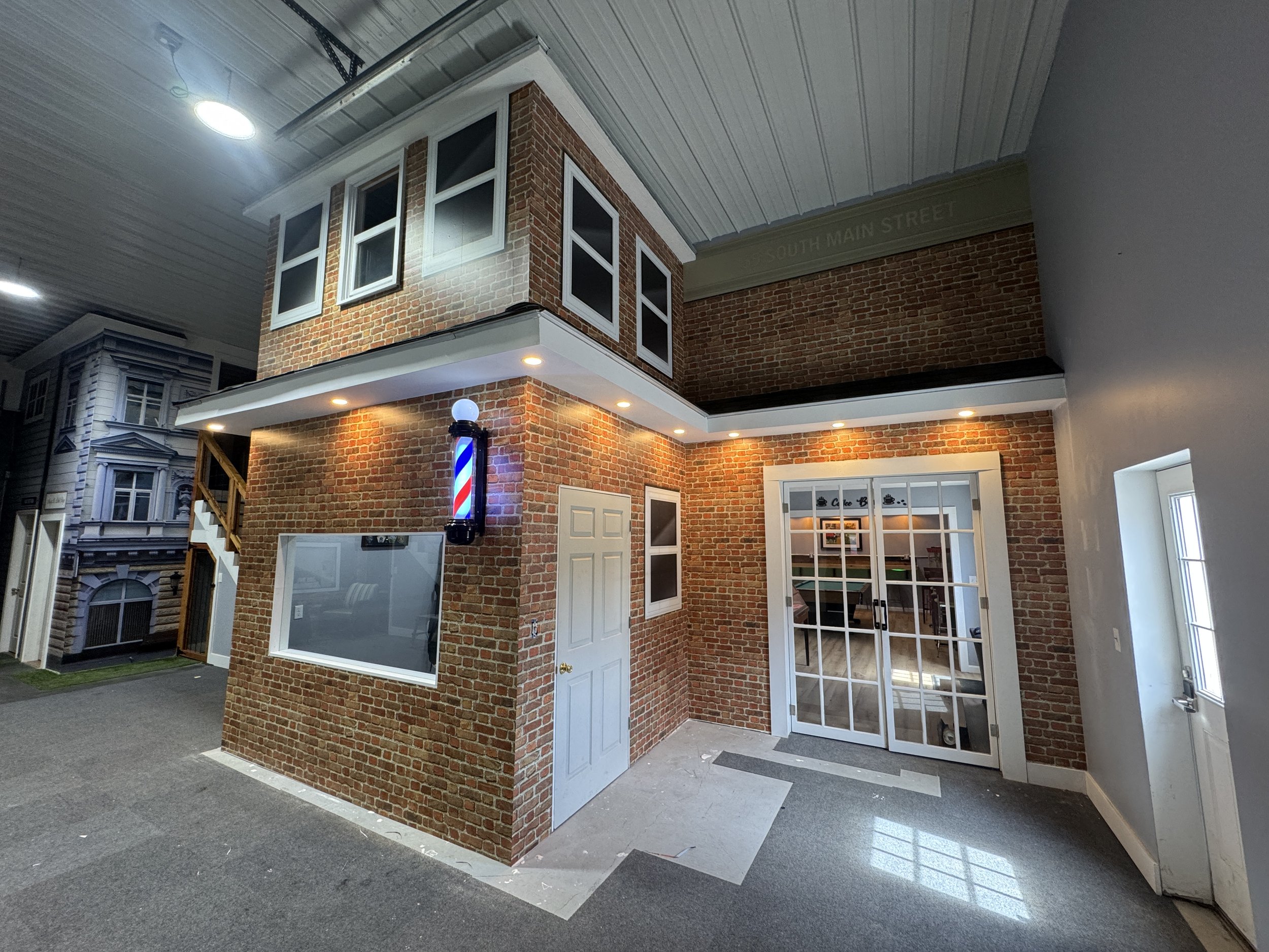 Indoor miniature house with brick exterior, multiple windows, a white door, and a corner with a barbershop pole. The room has a gray carpet and a gray ceiling with recessed lighting.