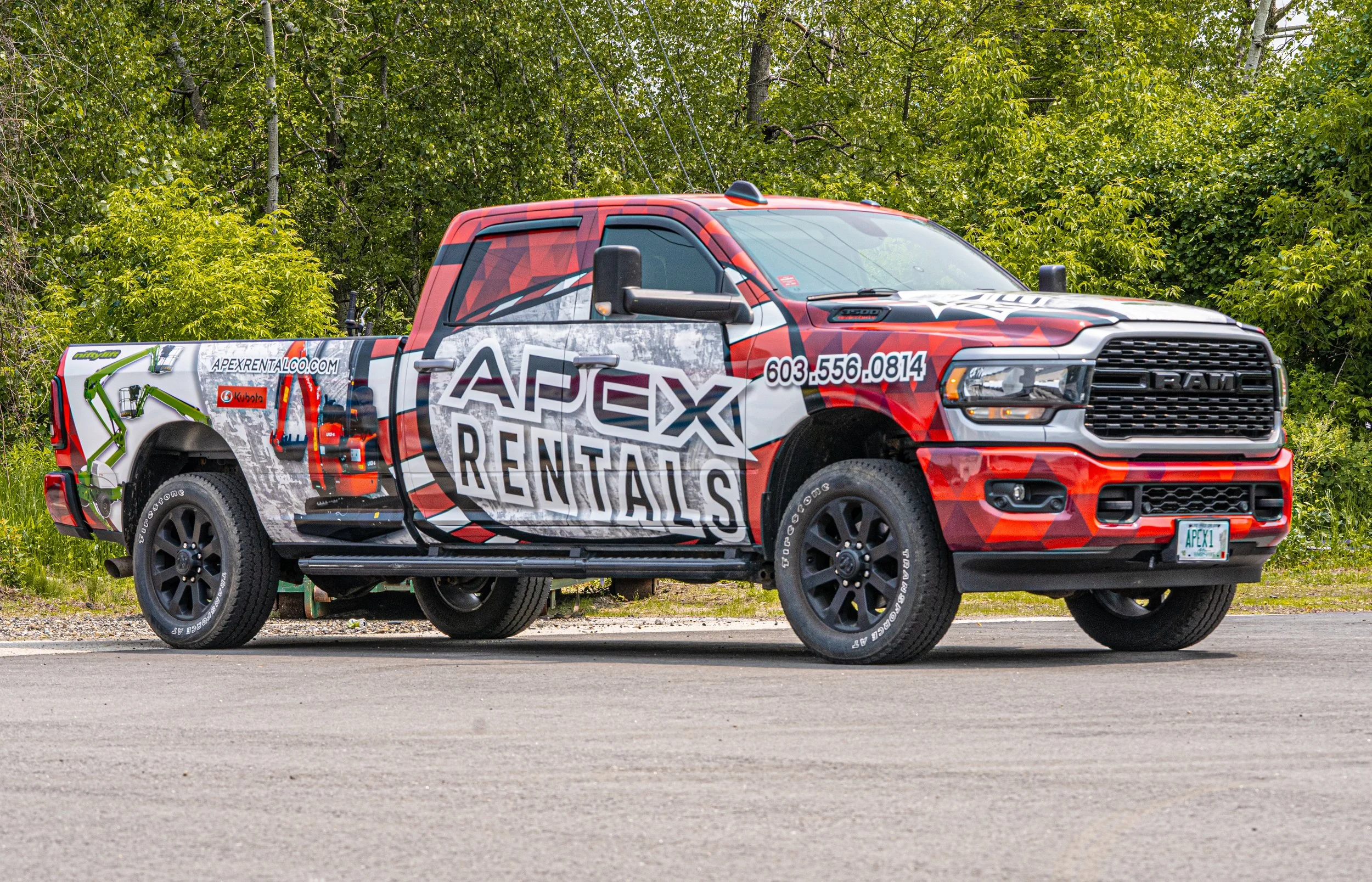 A pickup truck with branding for Apex Rentals, featuring a graphic of construction equipment, a phone number, and a website on the side, parked on a street with green trees in the background.