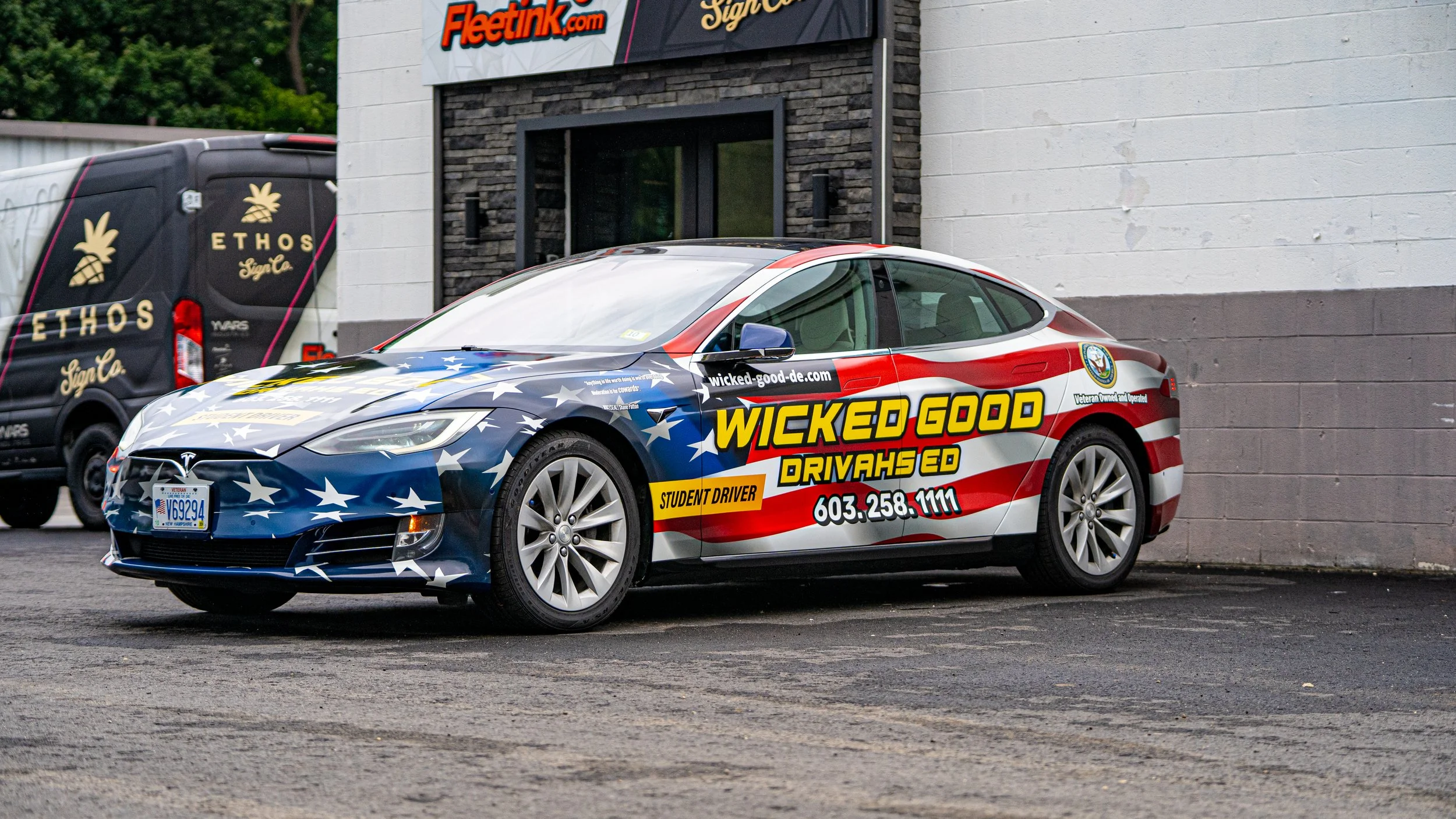 A Tesla electric car with a patriotic American flag design, advertising Wicked Good Drivahs ED, parked outside a building with a sign for Fleettink.com, and a black Ethos Style van nearby.
