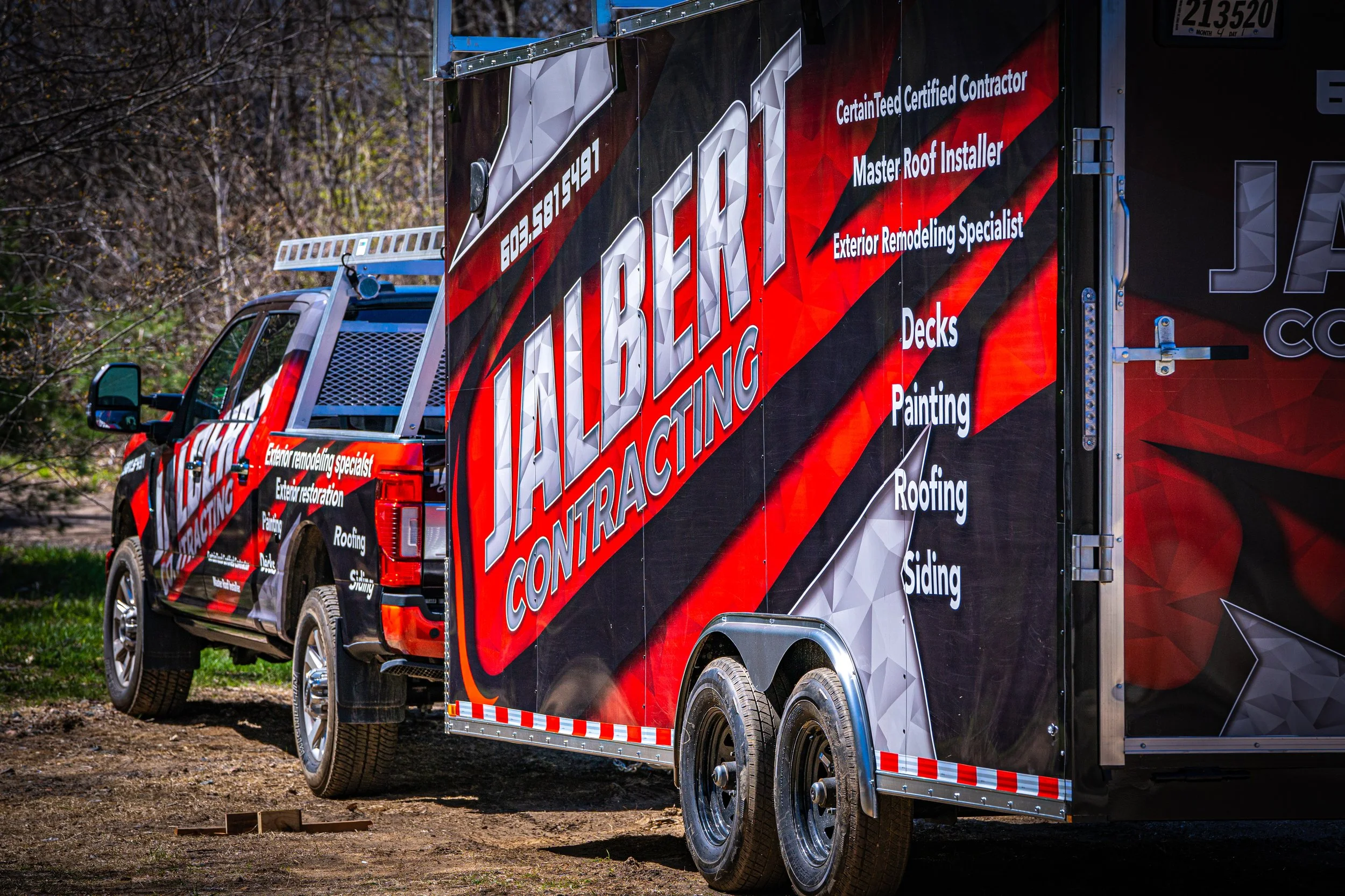 A red and black contractor truck with white text advertising roofing and exterior remodeling services parked outdoors near trees and dirt ground.