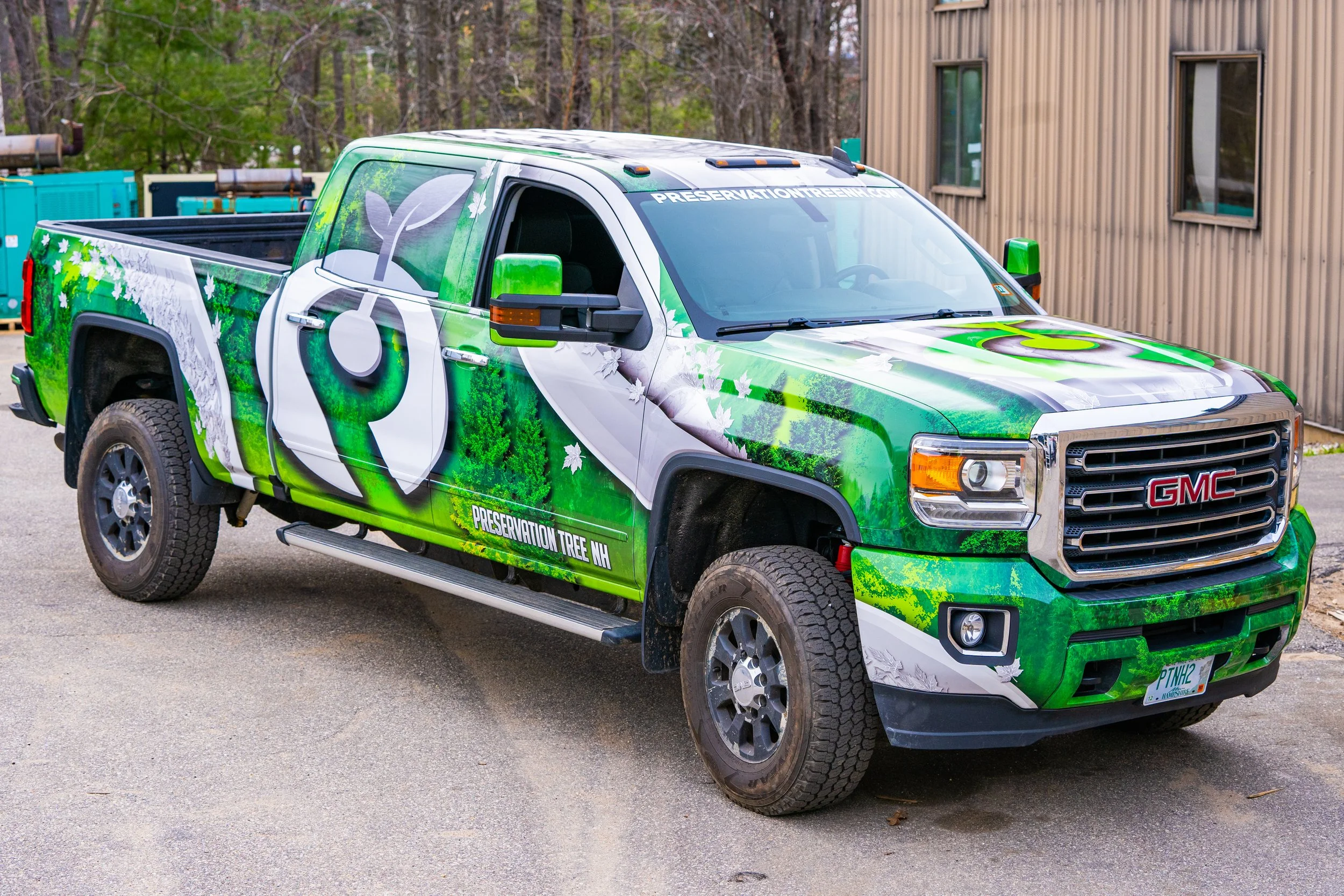 Green and white GMC pickup truck with tree and leaf graphics and the words PRESERVATION TREE NH on the side. The truck is parked outdoors near a wooden building with trees in the background.