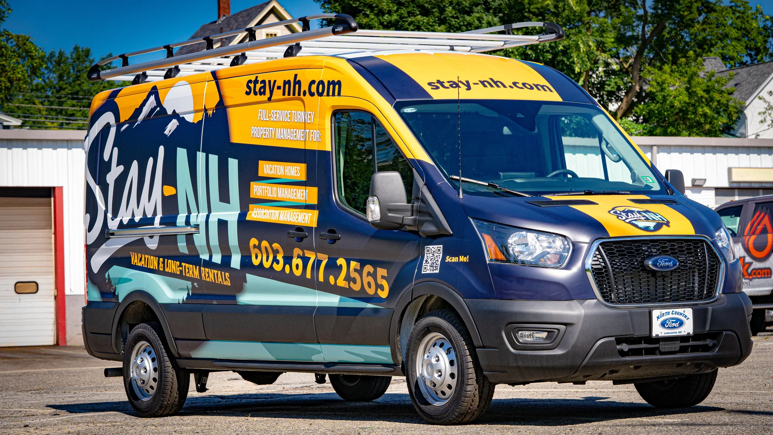Colorful company van with a mountainous design, advertising vacation rentals and property management services, parked outdoors with trees and buildings in the background.