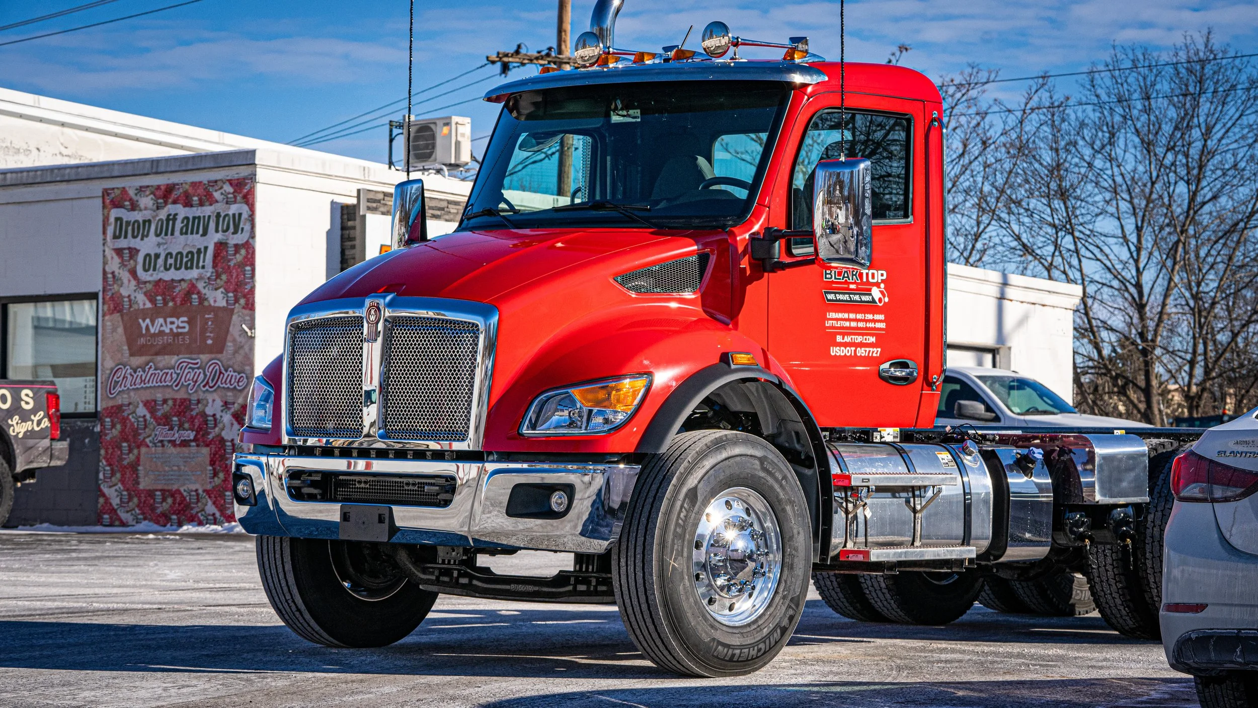 Red semi-truck parked in a lot with a white building in the background that has a sign reading 'Drop off any toy or coat!'
