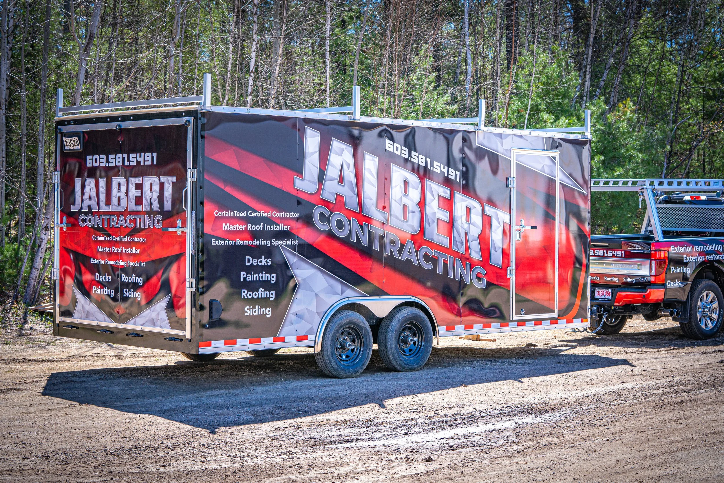 A mobile trailer for Jalbert Contracting, featuring company branding and services like roofing, siding, and painting, parked on a dirt area with a wooded background.