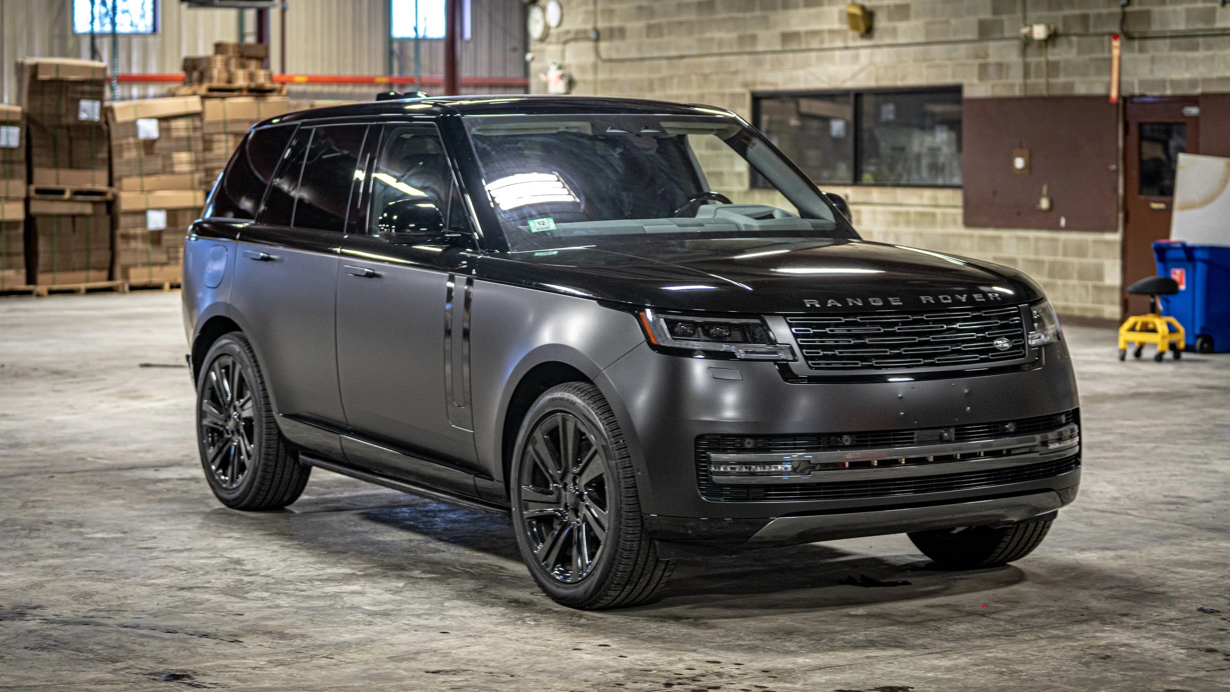 A black and gray Range Rover SUV parked inside a warehouse with stacks of boxes and equipment in the background.
