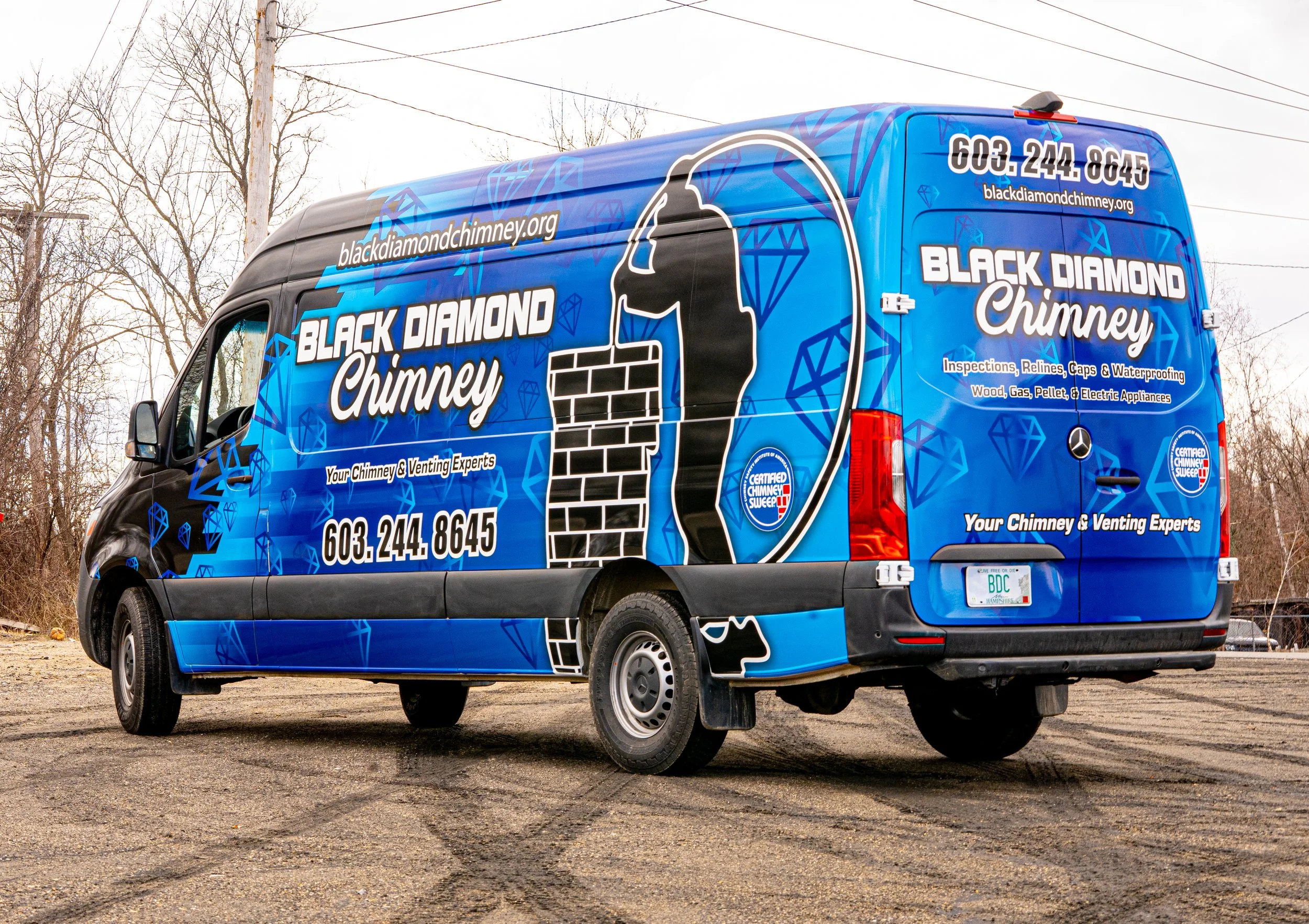A blue van with advertisements for Black Diamond Chimney, featuring a logo of a person laying bricks. The text includes the company's website, phone number, and services, with a background of blue diamond patterns. The van also has the phrase "Your Chimney & Venting Experts" and a Certified Chimney Sweep badge. The license plate reads "BDC." The van is parked on a gravel surface with leafless trees and power lines in the background.