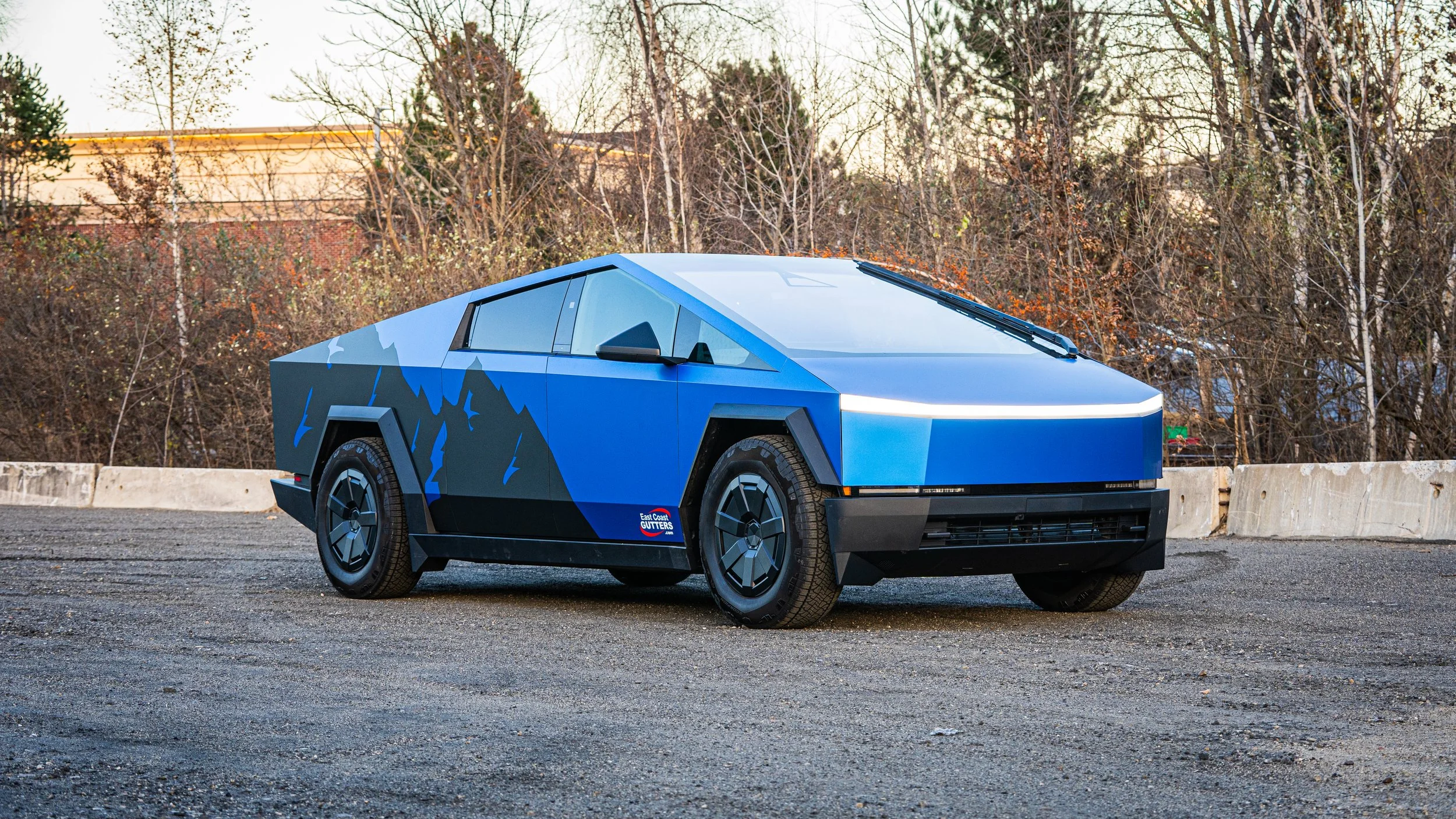 A futuristic electric blue vehicle with angular design and mountain decals parked on a paved road, with leafless trees and a beige building in the background.