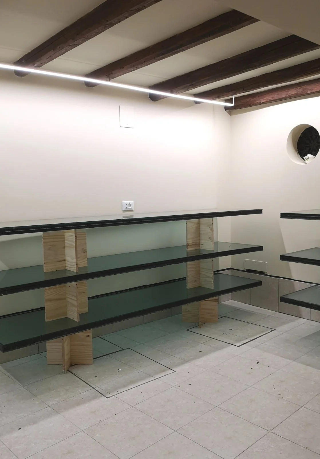 Interno di un locale vuoto con scaffali in vetro e legno, pavimento piastrellato e travi in legno a vista.
