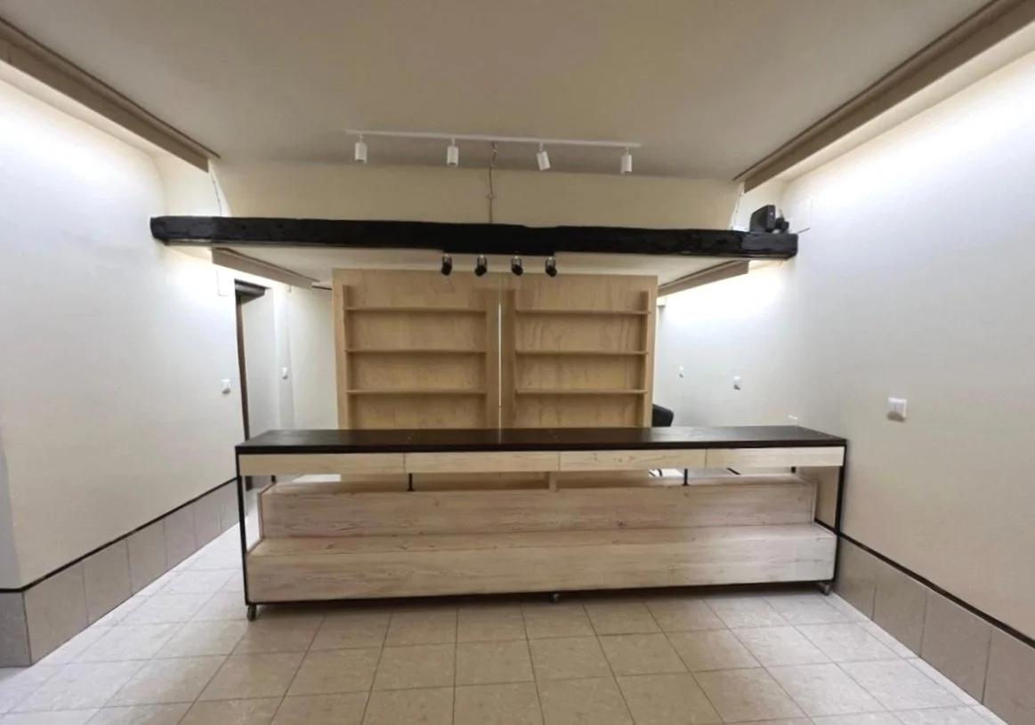 Interno di un negozio vuoto con bancone in legno, scaffalature posteriori e illuminazione a faretti.
