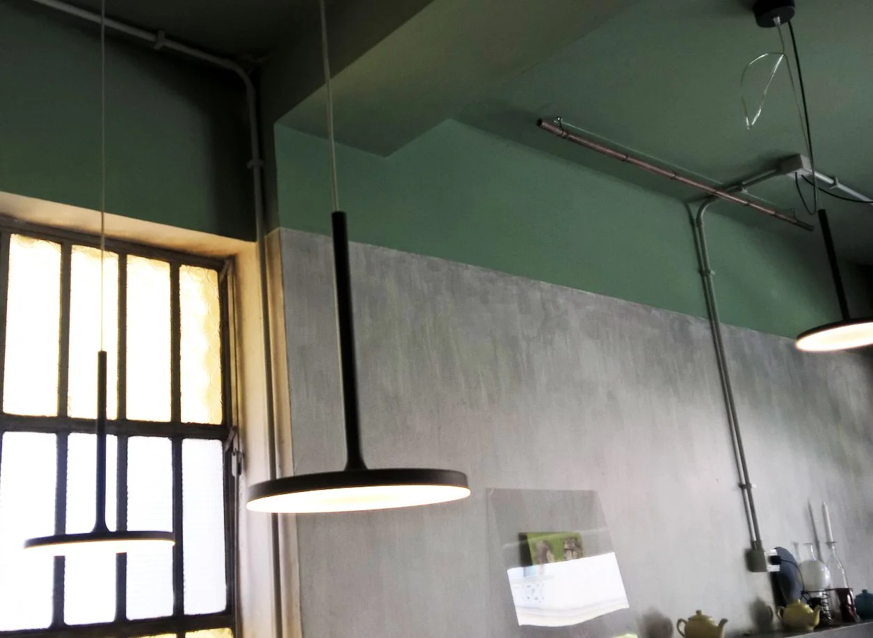 Interno di un locale con lampade a sospensione, finestra industriale, pareti verde e grigie e tubature a vista.
