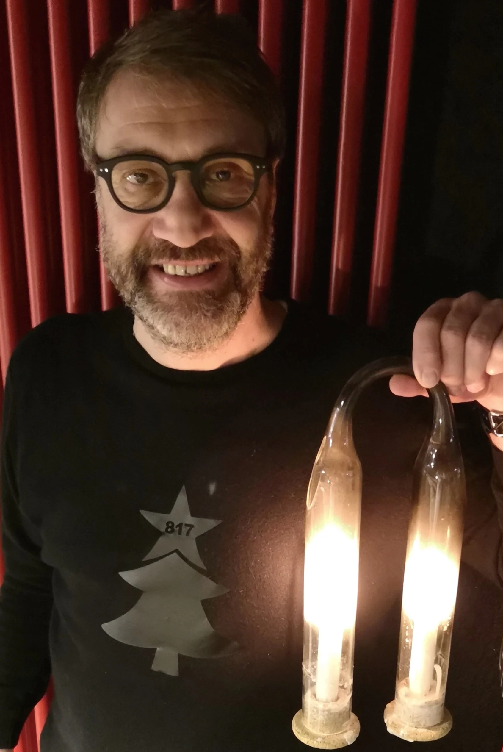 Uomo sorridente con occhiali e barba, davanti a tende rosse, accanto a due candele accese.