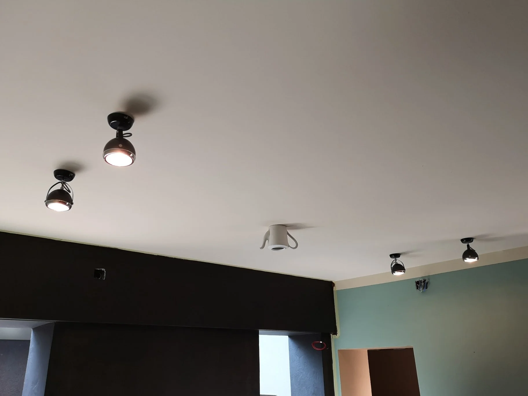 Soffitto interno con faretti neri accesi e un faretto bianco, sopra pareti scure e verde chiaro.