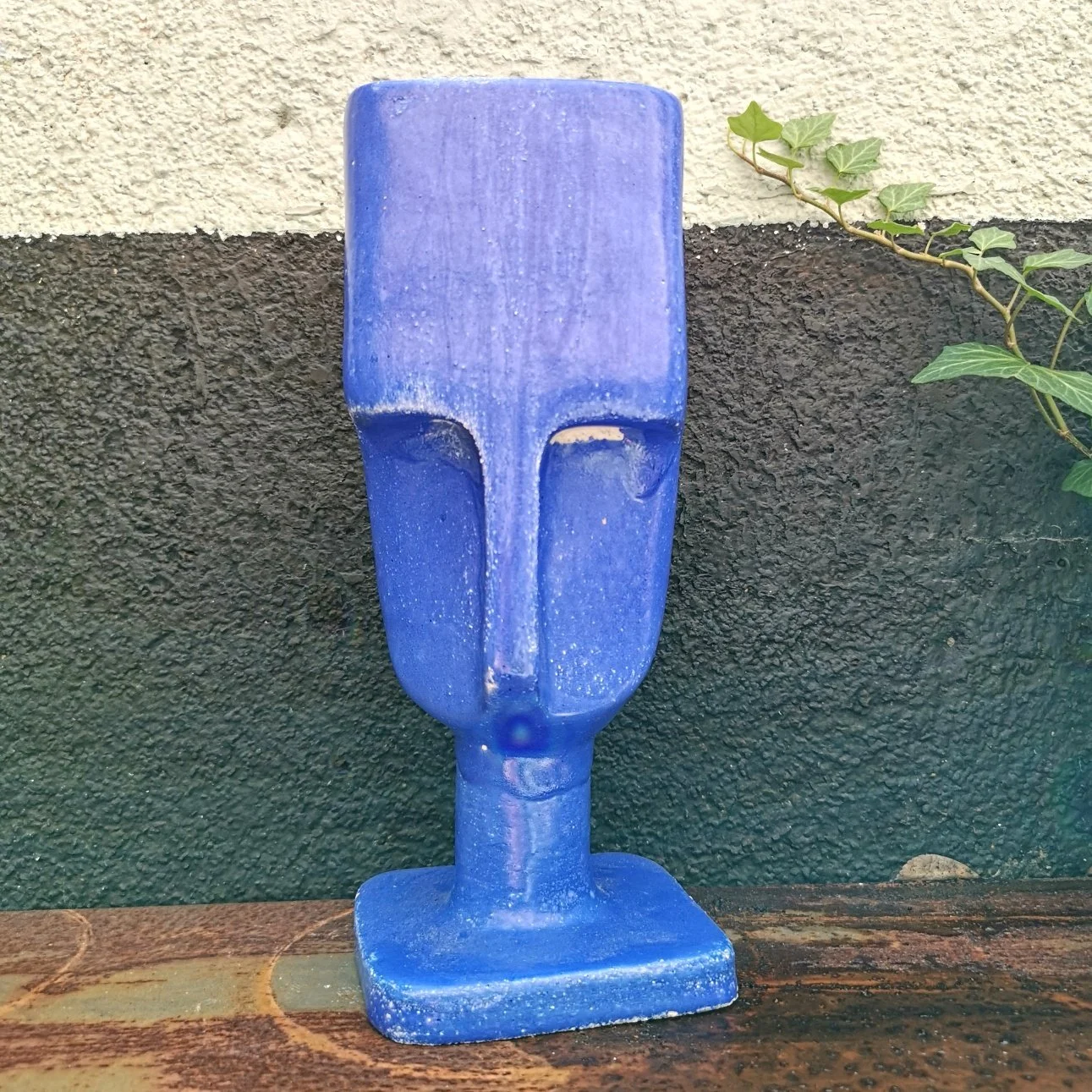 Scultura decorativa blu raffigurante un volto stilizzato, appoggiata su una base davanti a una parete con edera.