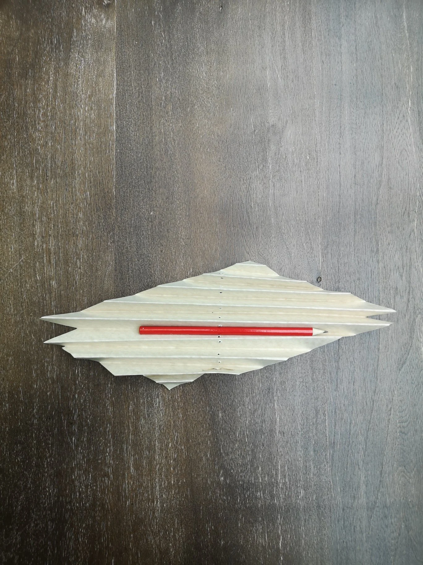 Matita rossa appoggiata su un foglio di carta piegato a forma di barca, su un tavolo di legno scuro.