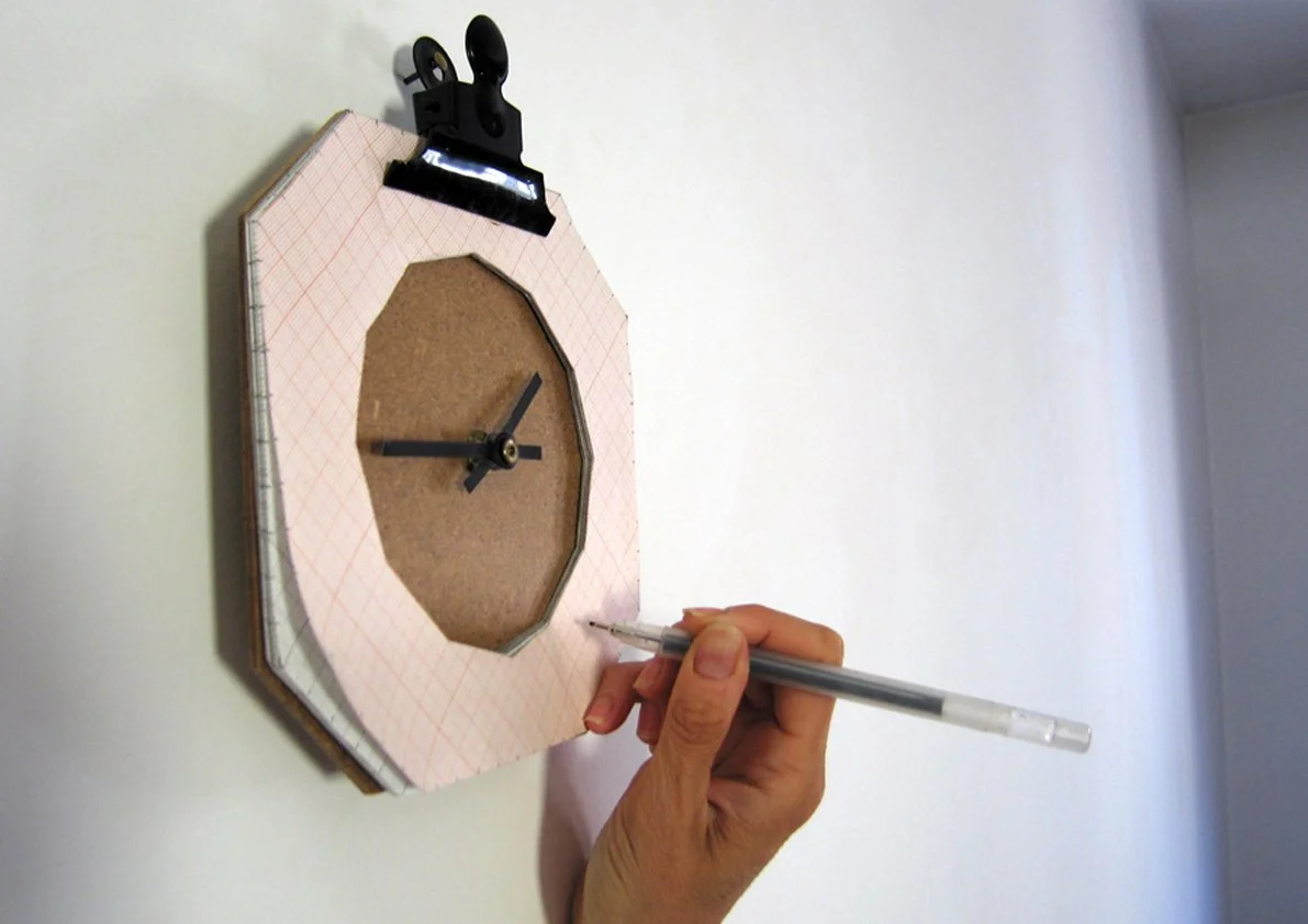 Mano che disegna con una penna su un modello di orologio artigianale fissato con una pinza.