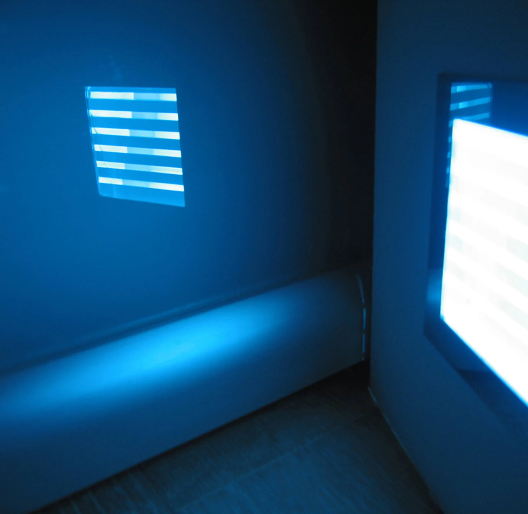 Angolo di una stanza immerso nella luce blu, con due finestre che proiettano riflessi di persiane sulle pareti e sul pavimento.