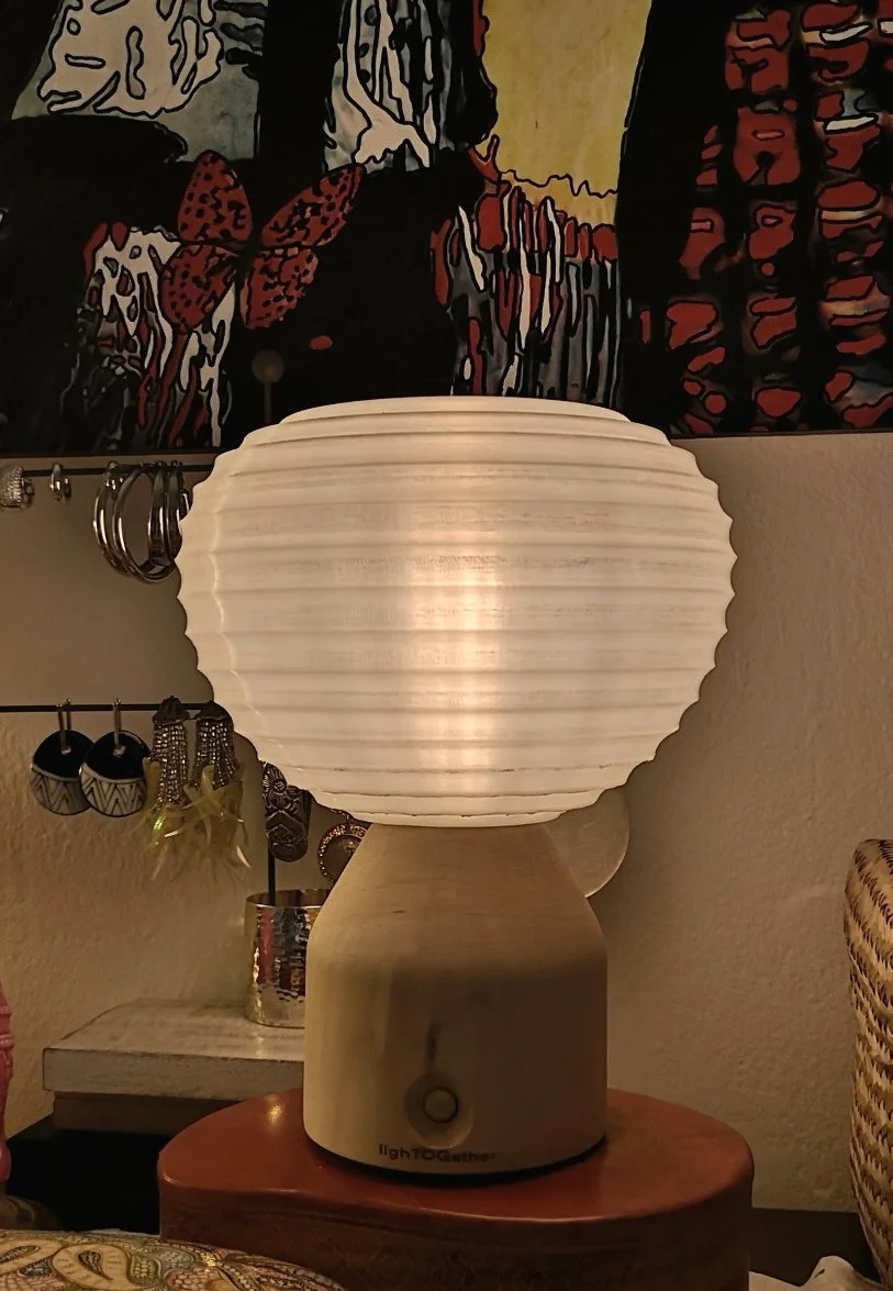 Lampada da tavolo bianca accesa su un mobile, con gioielli e opere d'arte colorate sullo sfondo.