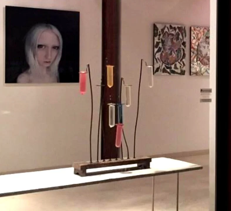 Installazione artistica con provette colorate sospese su una base, esposta in una galleria d’arte.