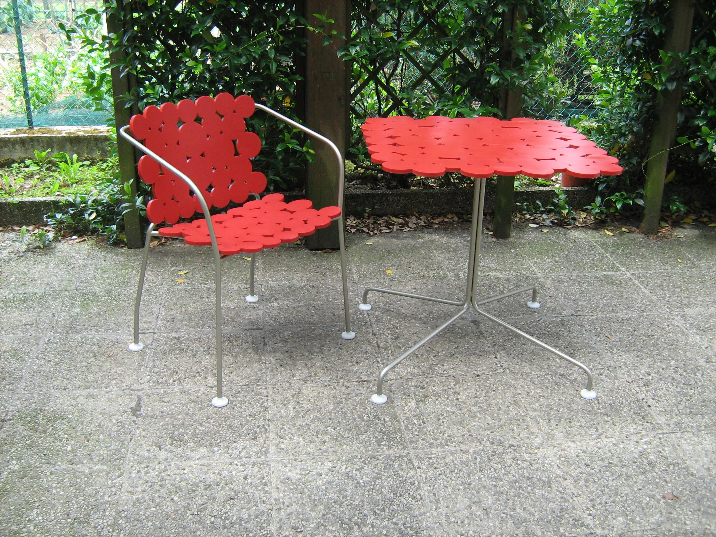 Tavolo da giardino rosso con due sedie decorate da motivi floreali, circondato da vegetazione.