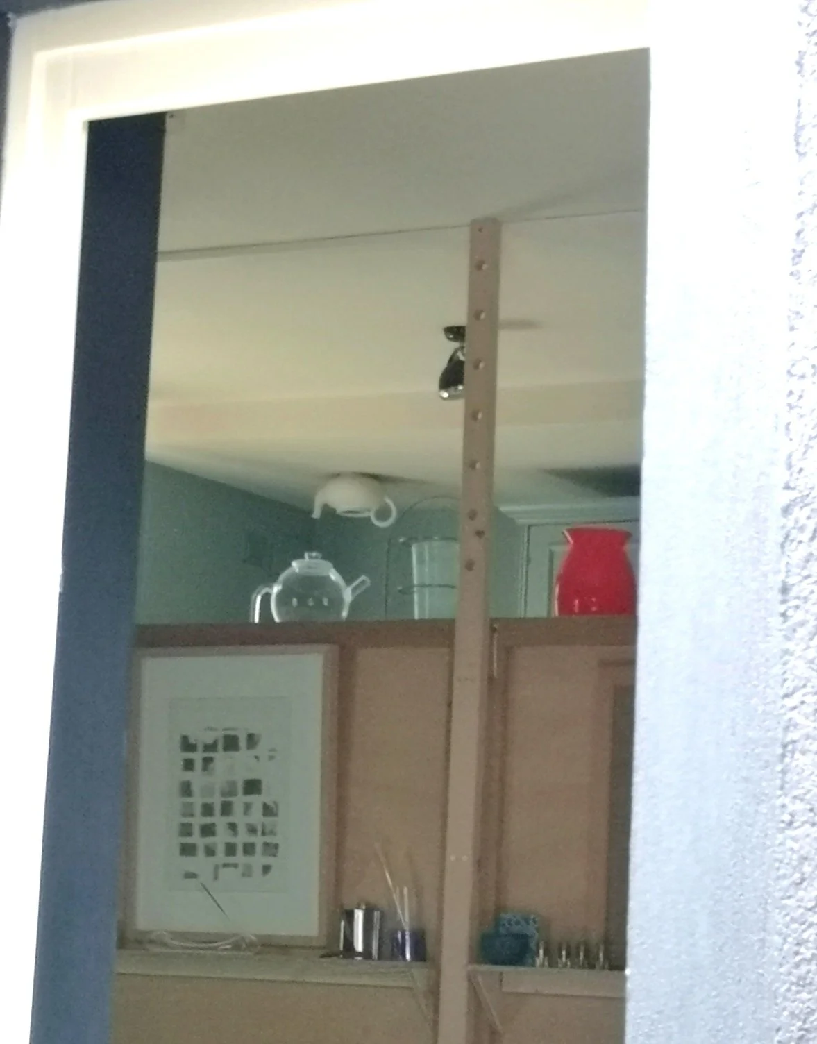 Specchio riflettente una cucina con credenza, teiera, bicchieri, brocca rossa e oggetti decorativi.