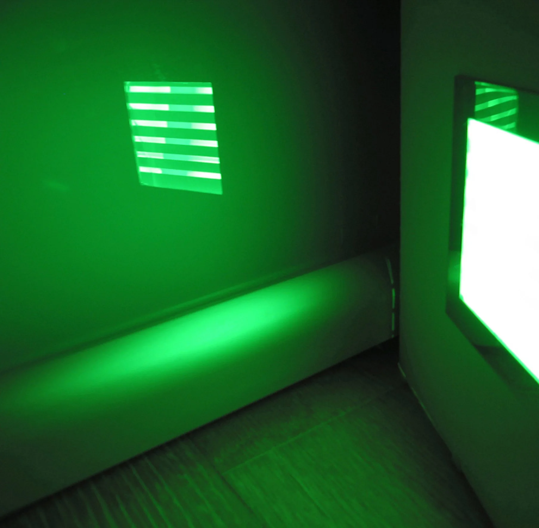 Stanza buia illuminata da luce verde, con pannello a strisce luminose sulla parete e grande schermo riflettente sulla destra.