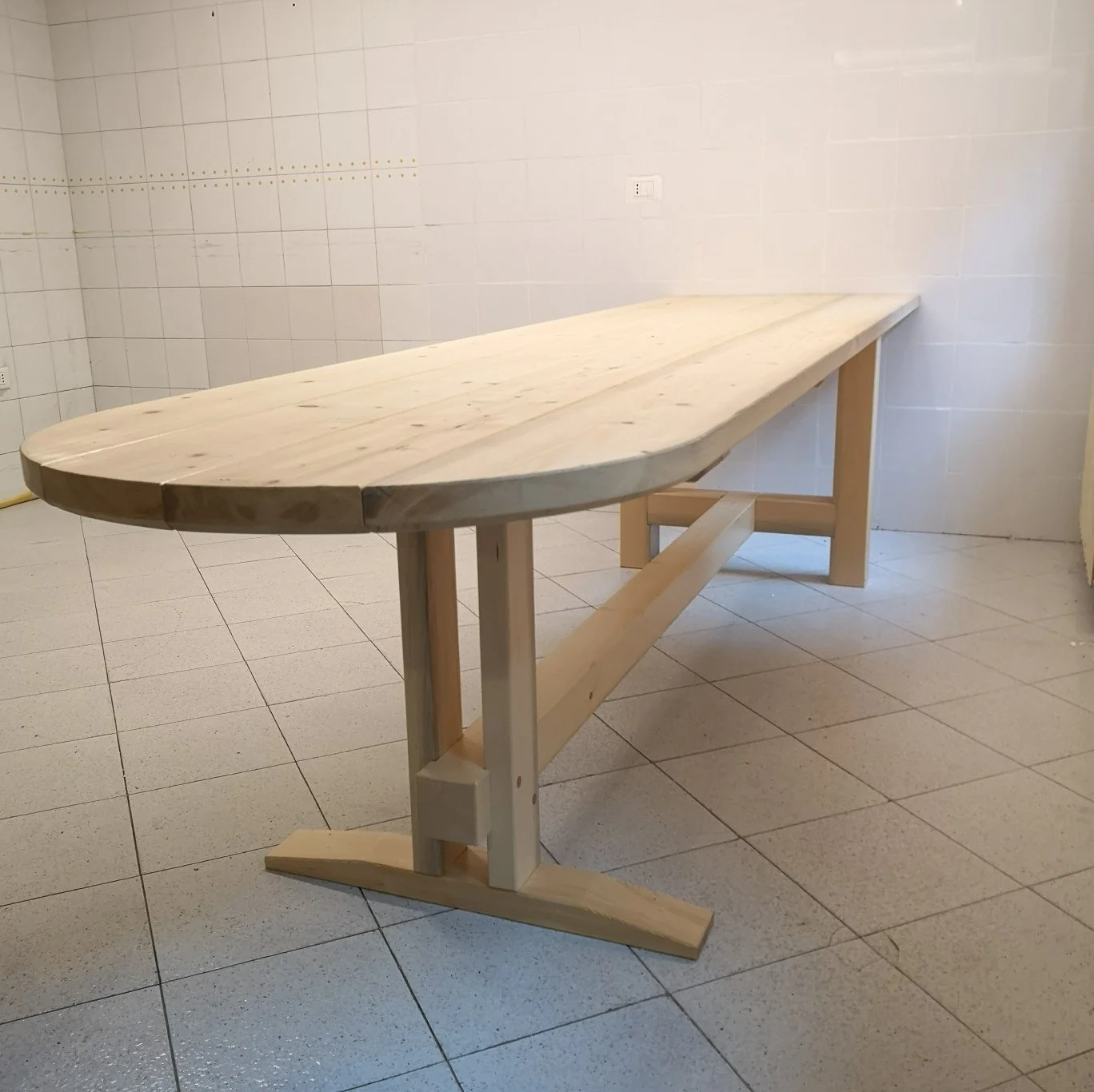 Grande tavolo da lavoro in legno chiaro con gambe e supporti rinforzati, in una stanza piastrellata