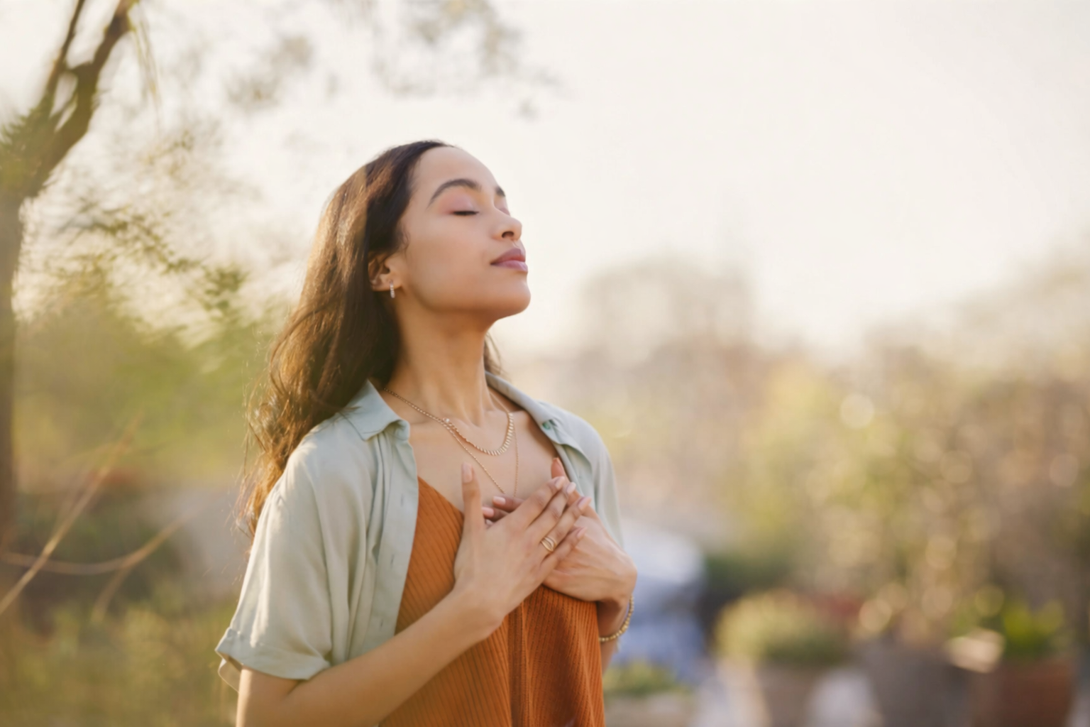 Young woman outdoors with eyes closed, hands on chest, enjoying peaceful moment.