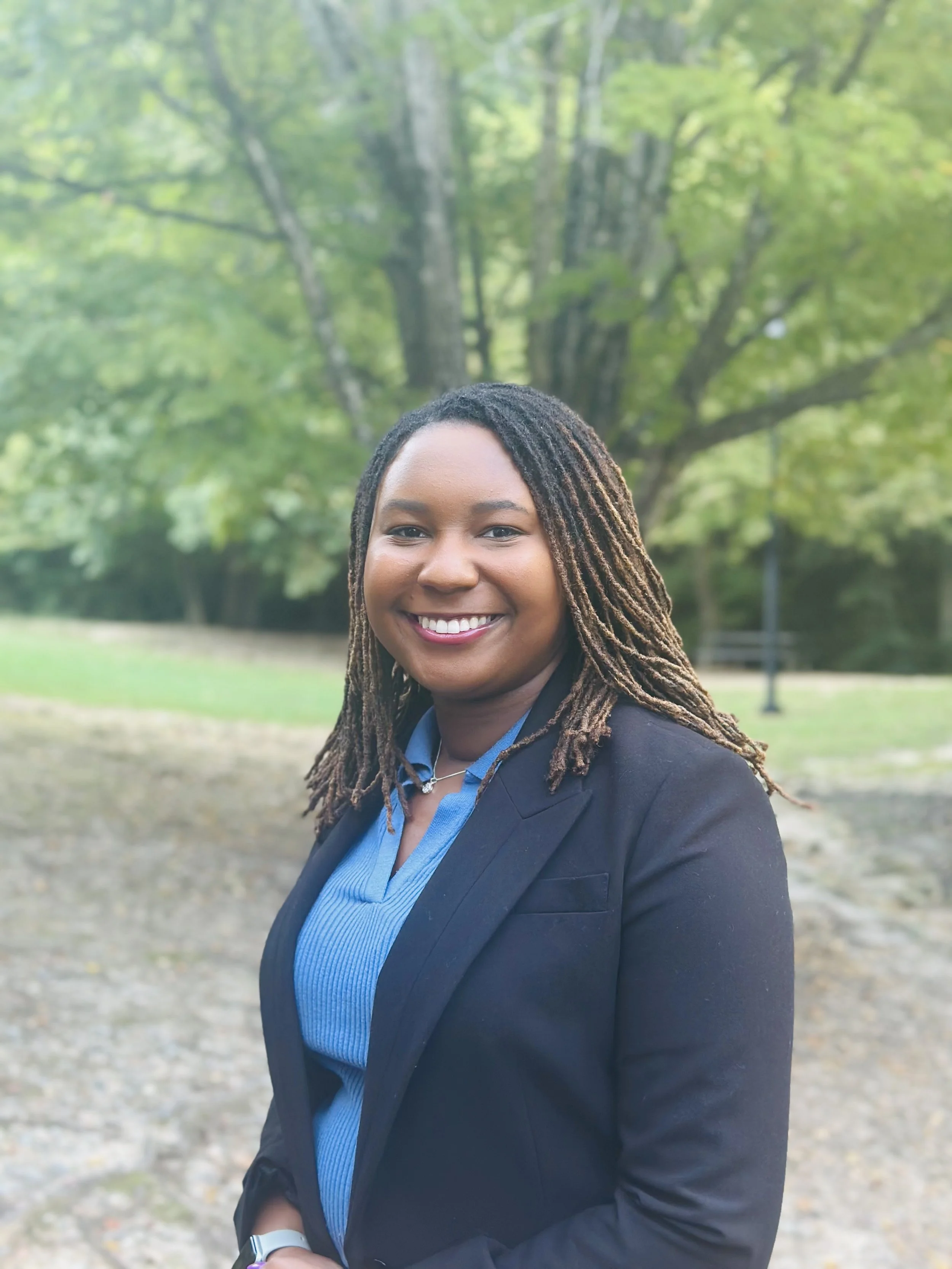 Smiling woman with dreadlocks wearing a black blazer over a blue shirt, standing outdoors in a park with a large green tree in the background.