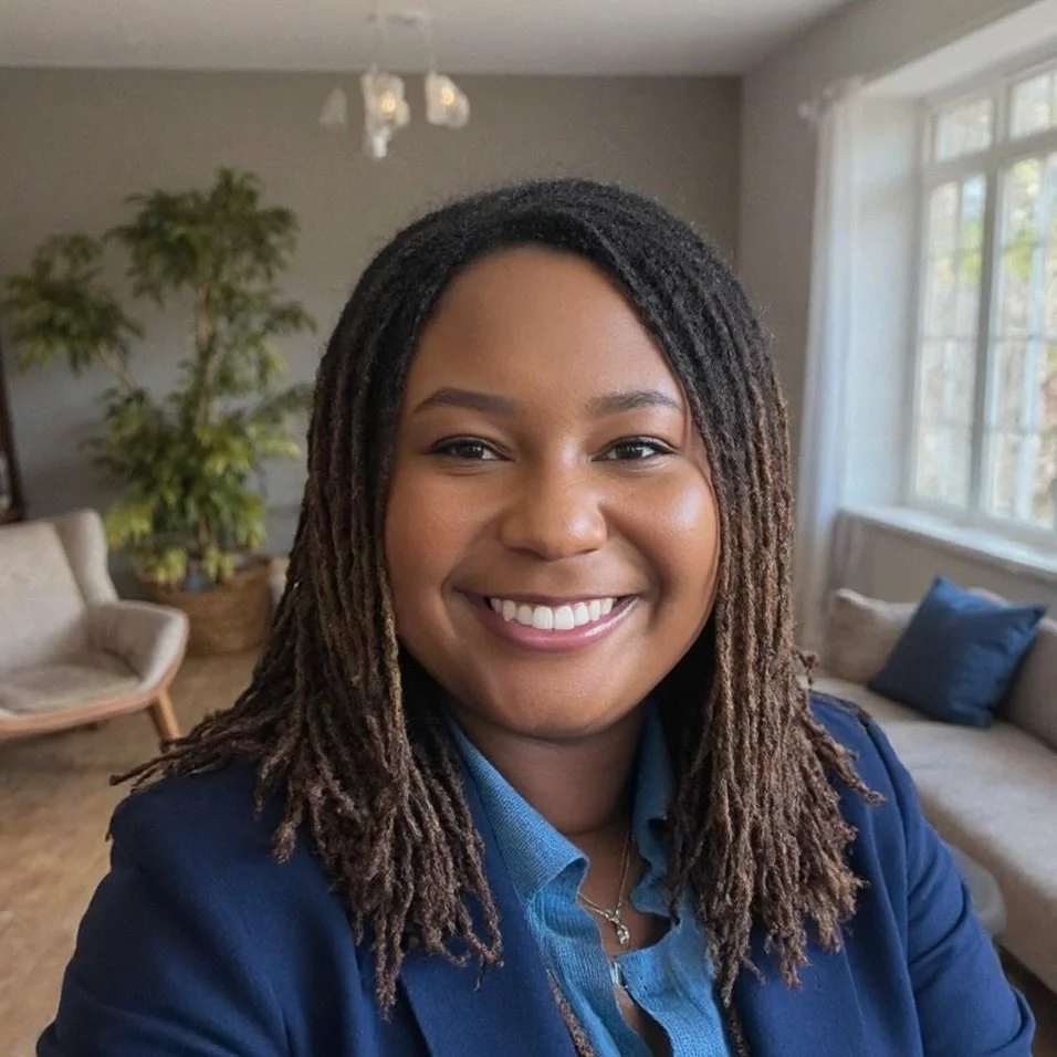 A smiling woman with dreadlocks wearing a blue blazer in a living room with a couch, pillows, a window, and a large houseplant.
