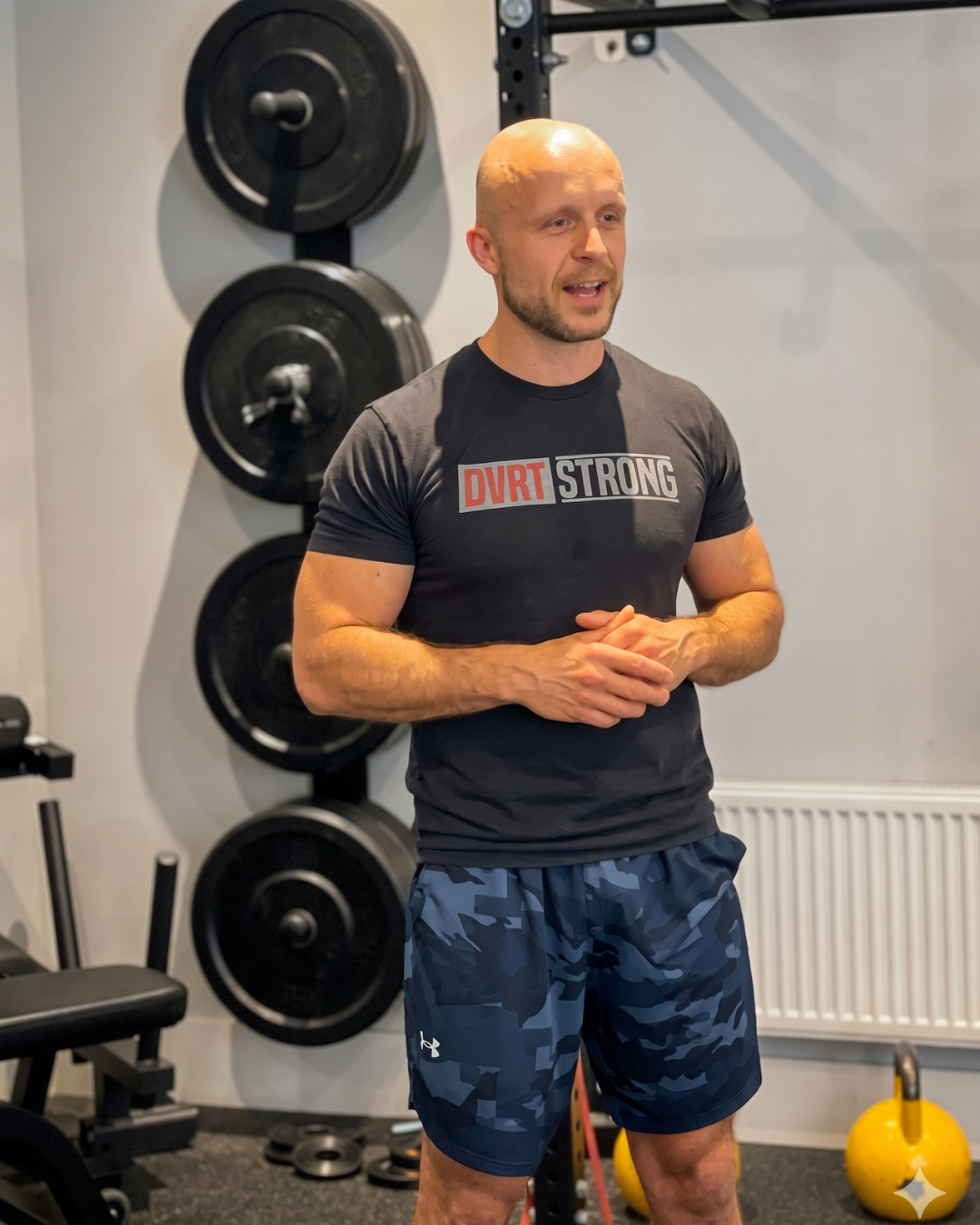 A muscular man with a bald head standing in a gym, wearing a black T-shirt with "DVRT STRONG" printed on it and blue camo shorts, speaking with his hands clasped together.