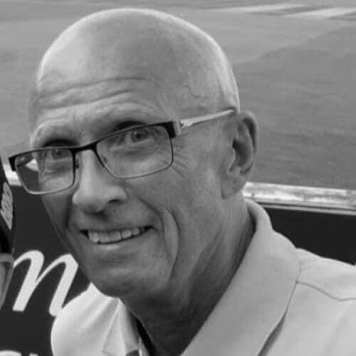 Black and white photo of a bald man wearing glasses, smiling, and wearing a collared shirt.