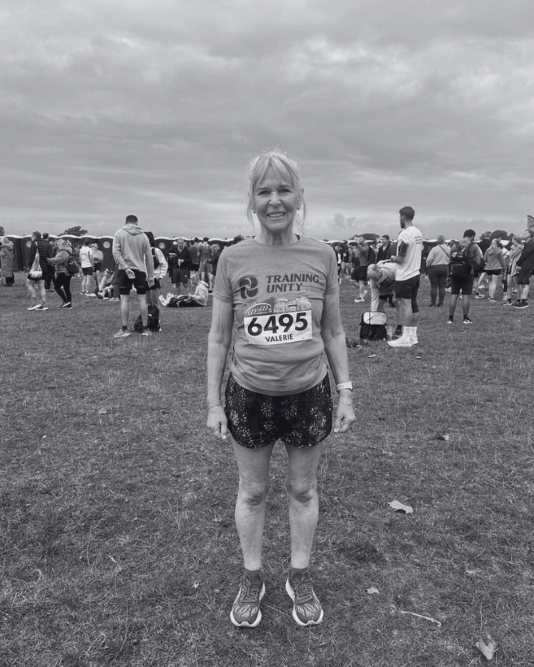 A woman standing outdoors at a race event with a crowd of runners behind her. She is wearing athletic clothing and a race bib with the number 6495 and the name Valerie.