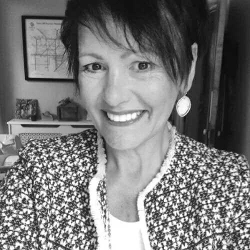 Smiling woman with short, dark hair and large earrings, wearing a patterned jacket, in an indoor setting.