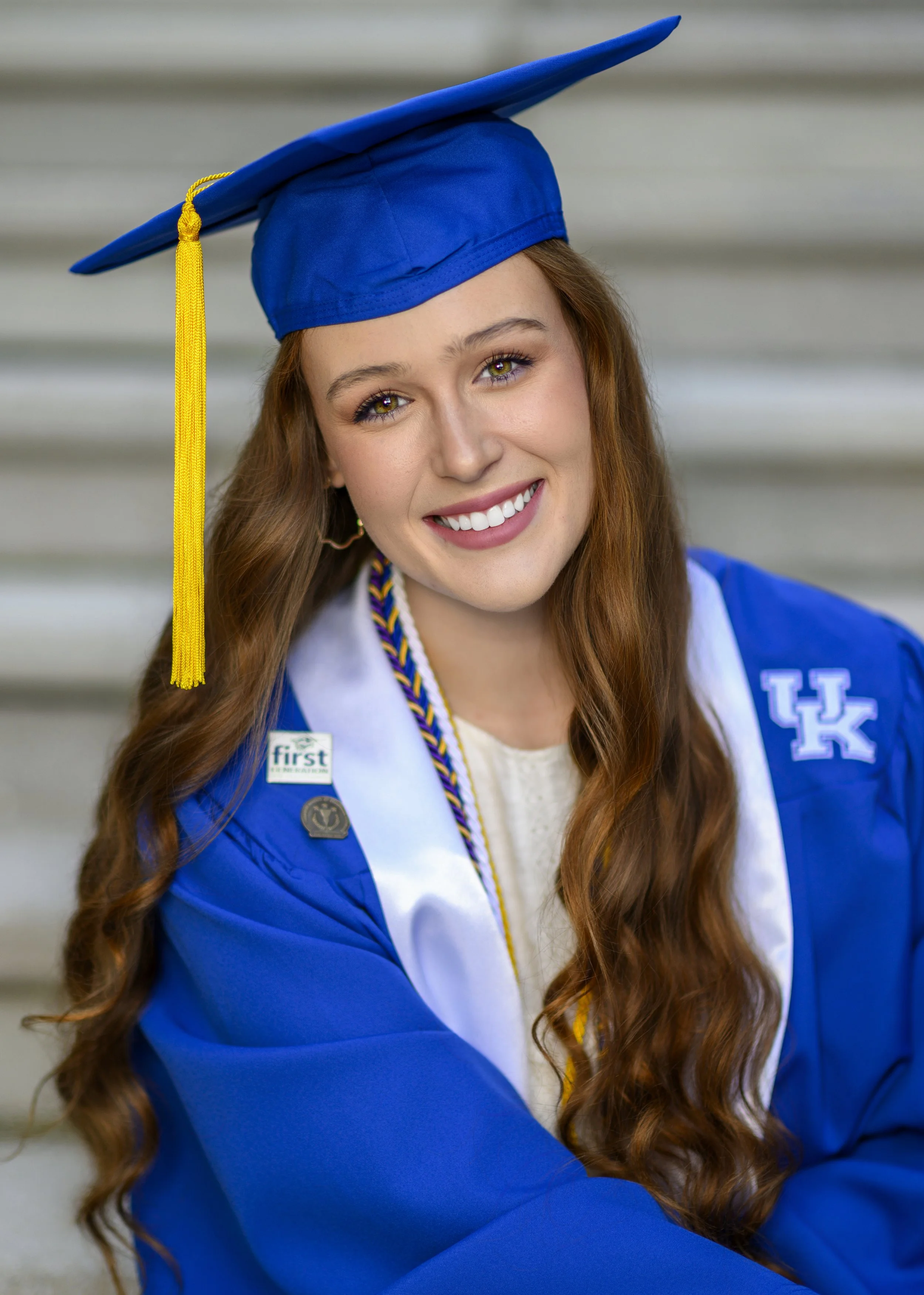 A young woman with long wavy red hair and green eyes wearing a blue graduation cap with a yellow tassel and a blue graduation gown, standing against a blurred outdoor background.