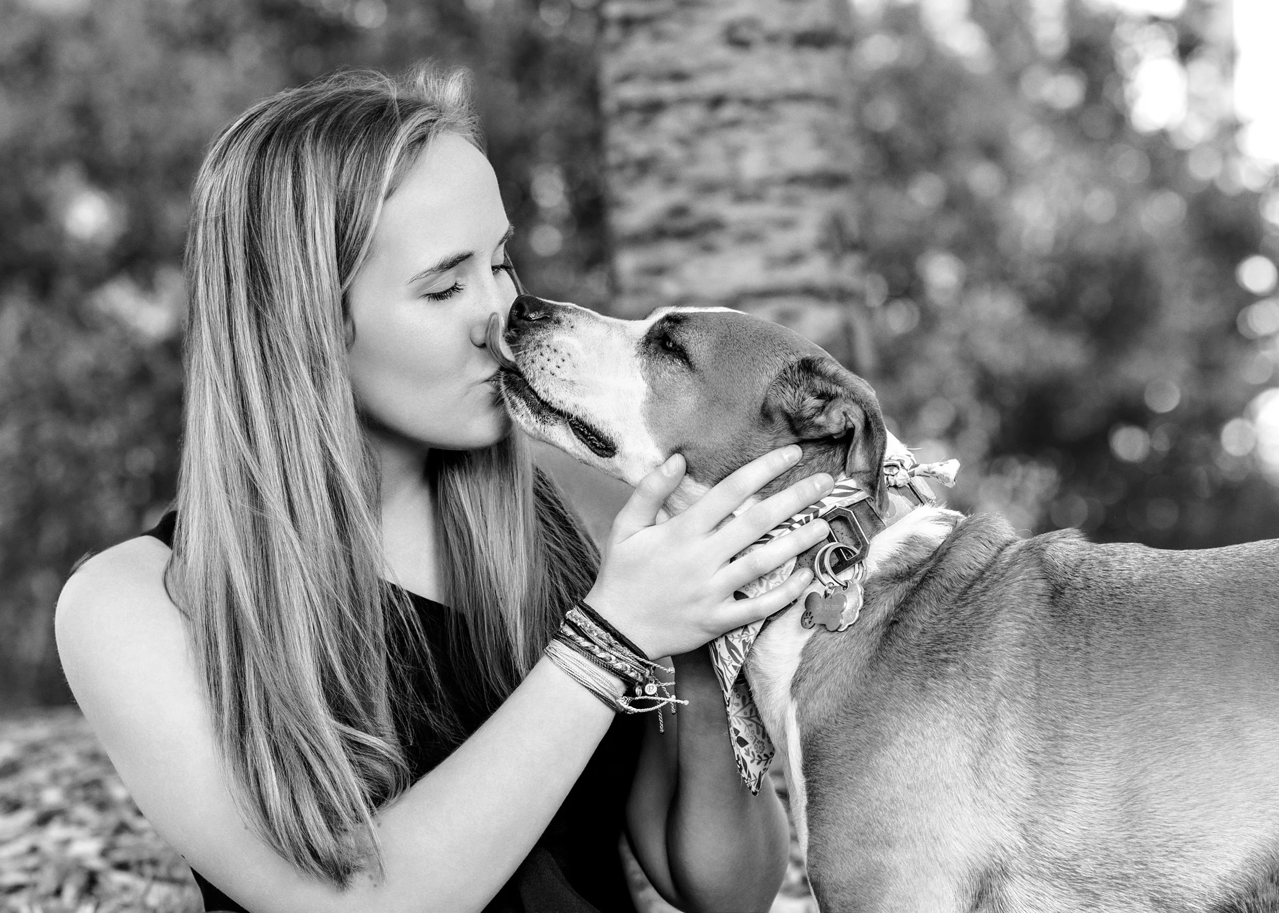 A young woman with long hair kissing a dog on the nose outdoors, with trees in the background.