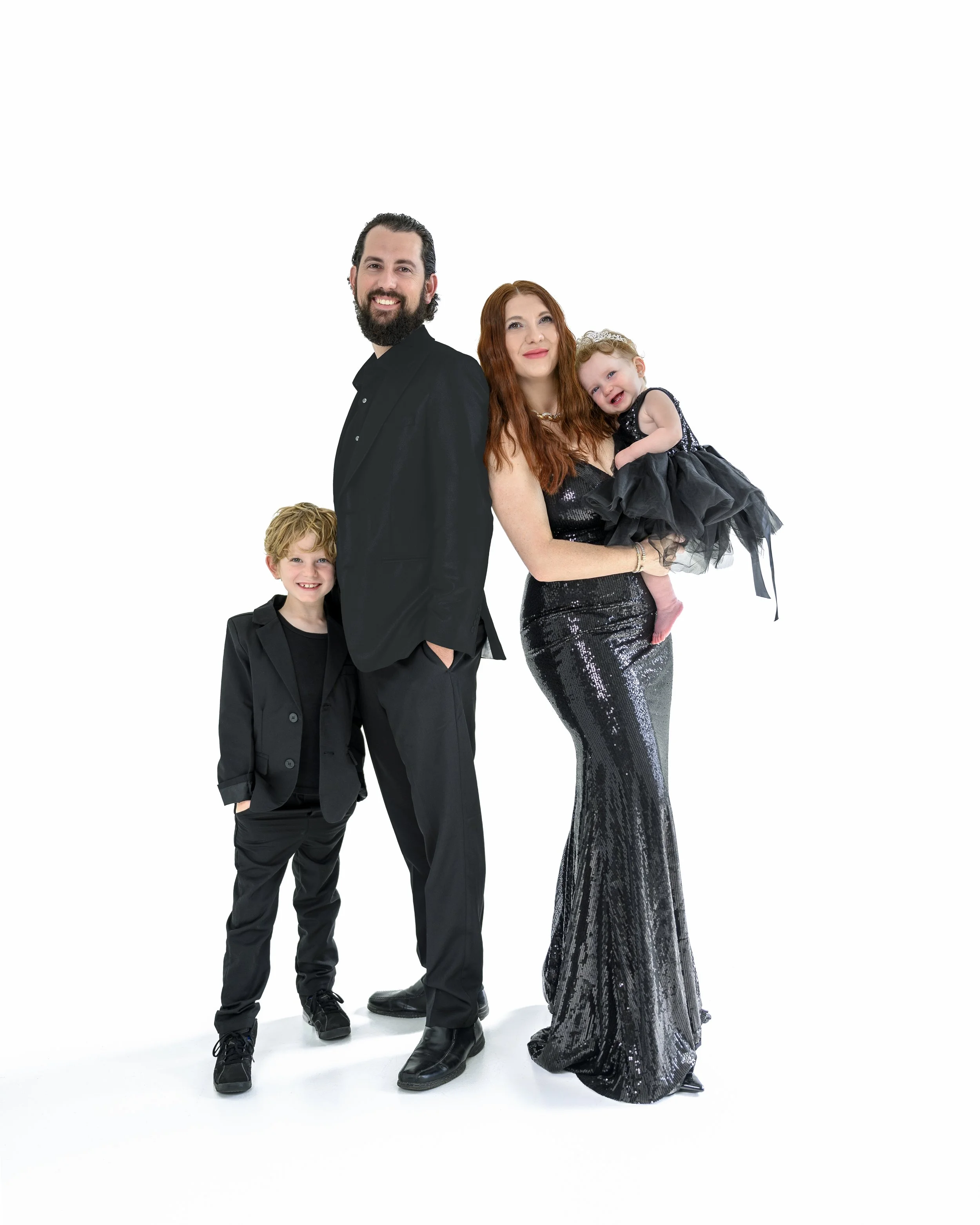 A multi-generational family of four dressed in black and silver outfits standing against a white background. The man has dark hair and a beard, wearing a black suit, and smiling. The woman has long red hair, wearing a shiny black dress, holding a you