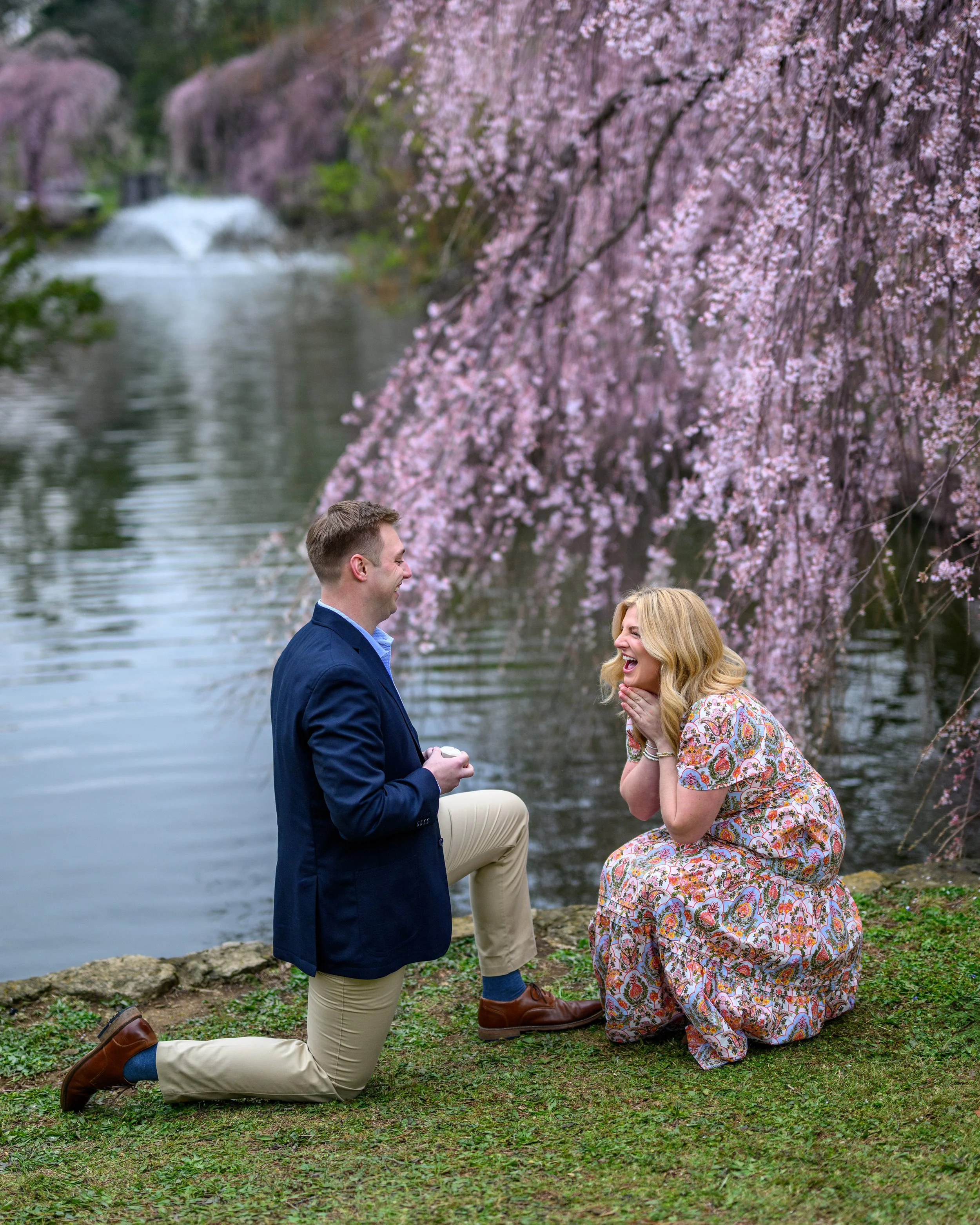 A man kneeling on one knee, holding a small box, proposing to a woman who is smiling and covering her mouth with her hands, by a river with cherry blossom trees in bloom.