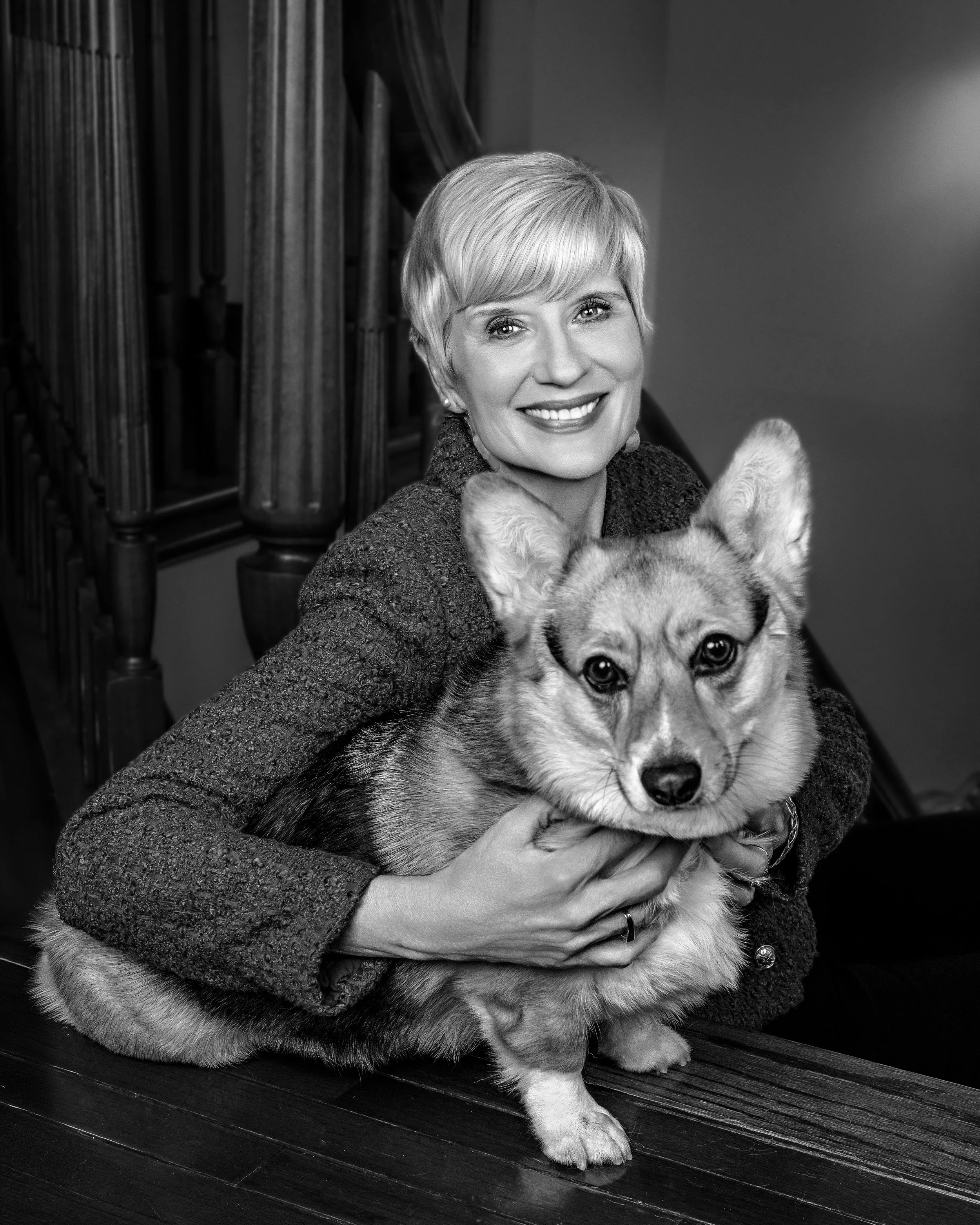 Black and white photo of a woman with short blonde hair smiling while holding a large dog, possibly a Husky, in front of a staircase made of wood.