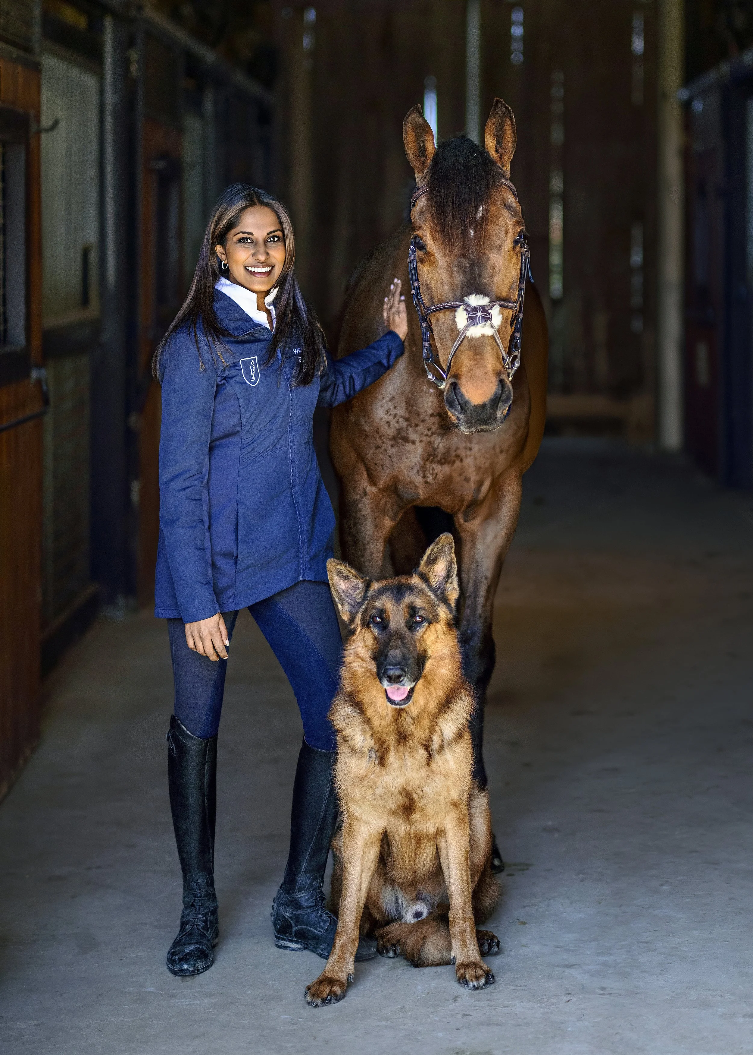 A woman in equestrian attire standing in a stable next to a brown horse and a German Shepherd dog. The woman is smiling and touching the horse.