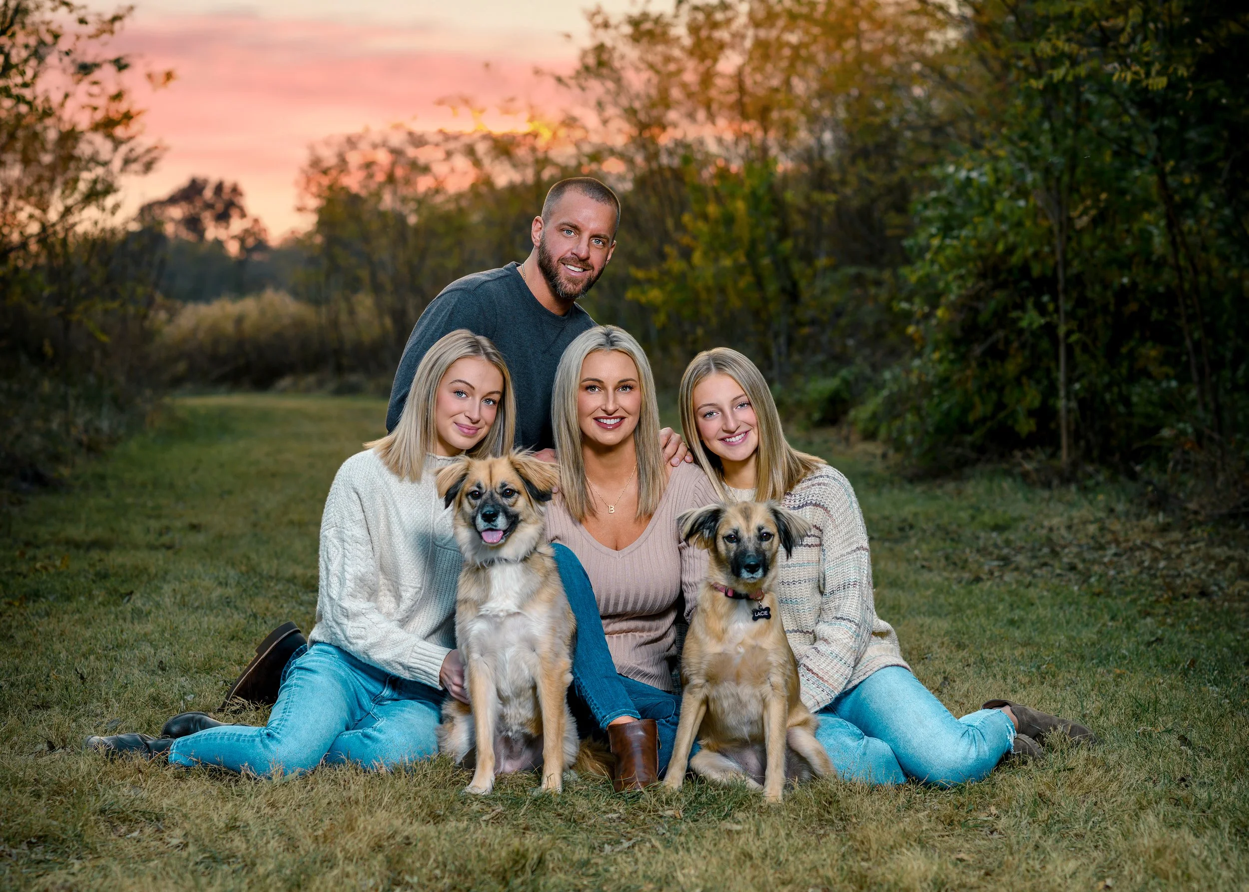 A group of four women and one man outdoors during sunset, sitting and kneeling on grass with two dogs, two young women on the left, a middle-aged woman in the center, a young woman on the right, and a man standing behind them, all smiling and wearing