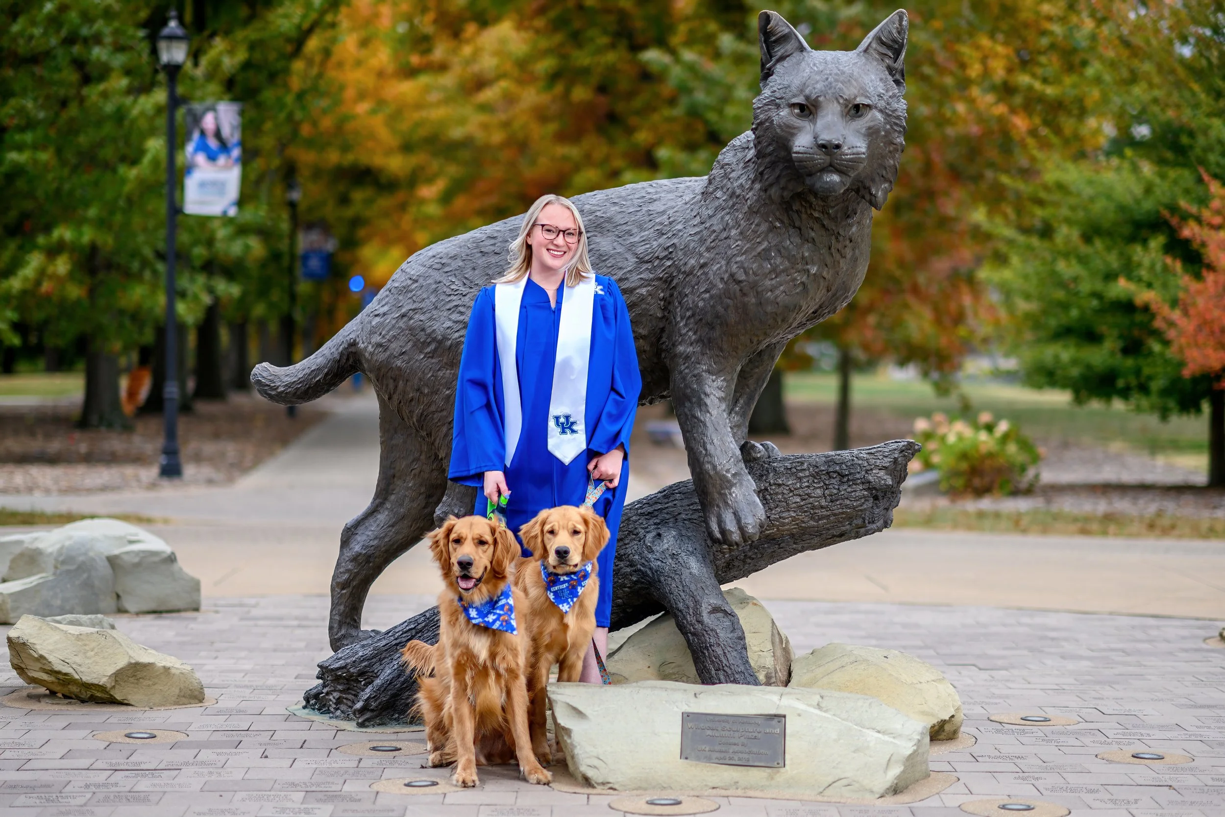 A woman in a blue graduation gown and white stole standing next to a large bronze statue of a wolf, with two golden retriever puppies wearing blue bandanas in front of her, in a park with autumn foliage.