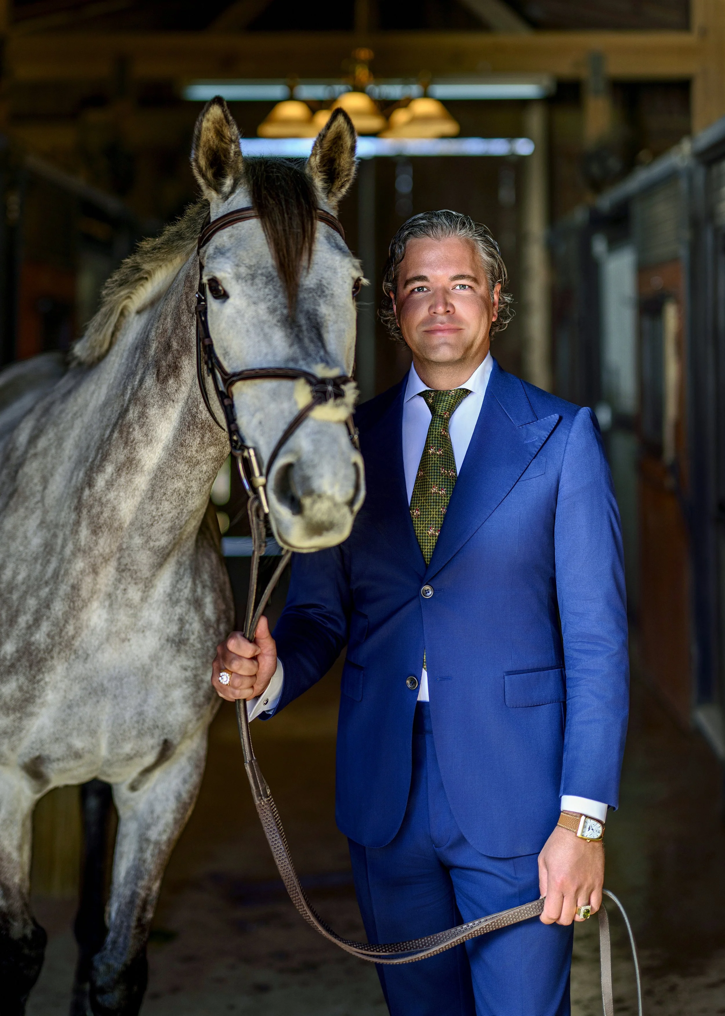 A man in a blue suit holding a gray horse's lead rope inside a stable.