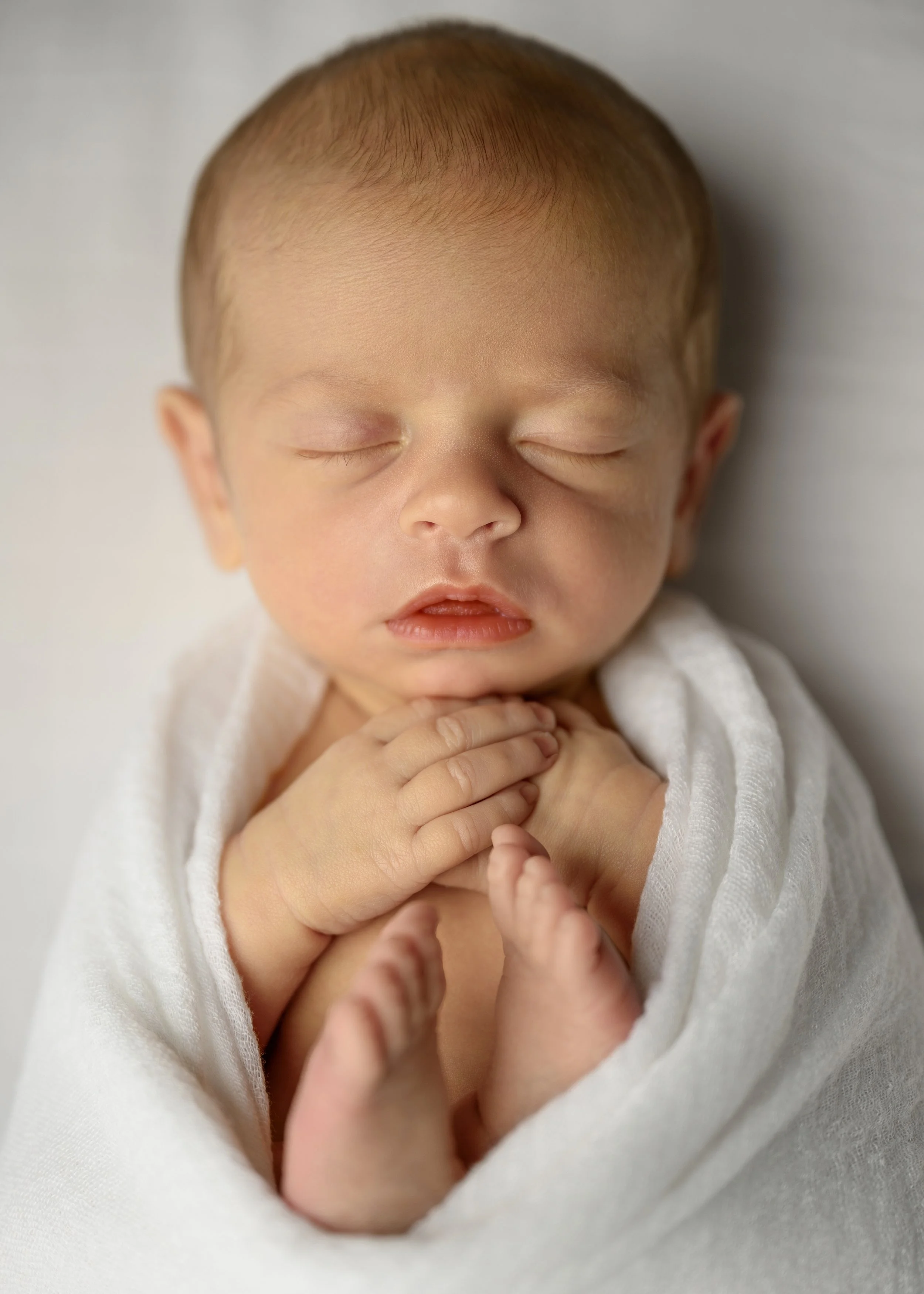 Close-up of a sleeping baby's face with hands clasped together, wrapped in a white blanket.