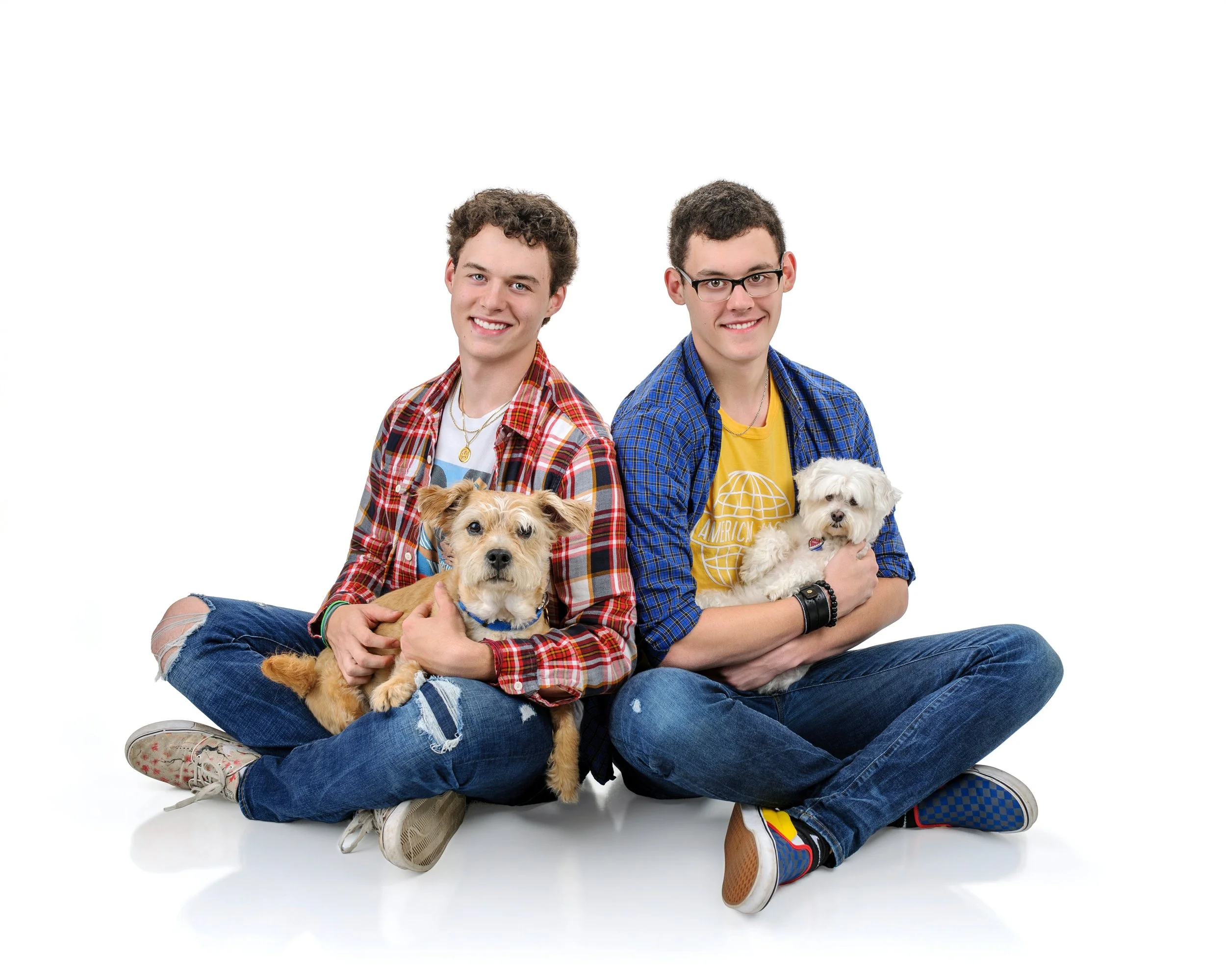 Two young men sitting cross-legged on the floor each holding a small dog, smiling, against a plain white background.
