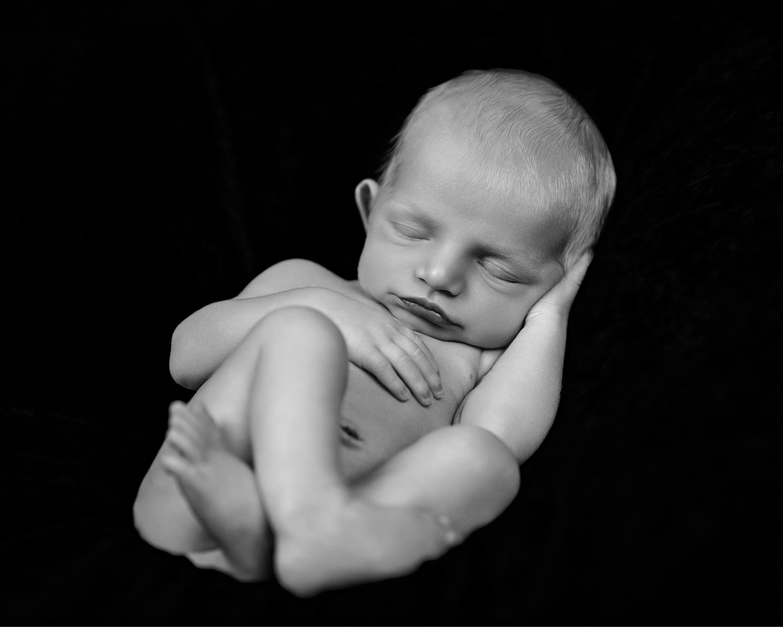 Black and white photo of a sleeping baby with short hair, lying on a soft surface with a dark background.
