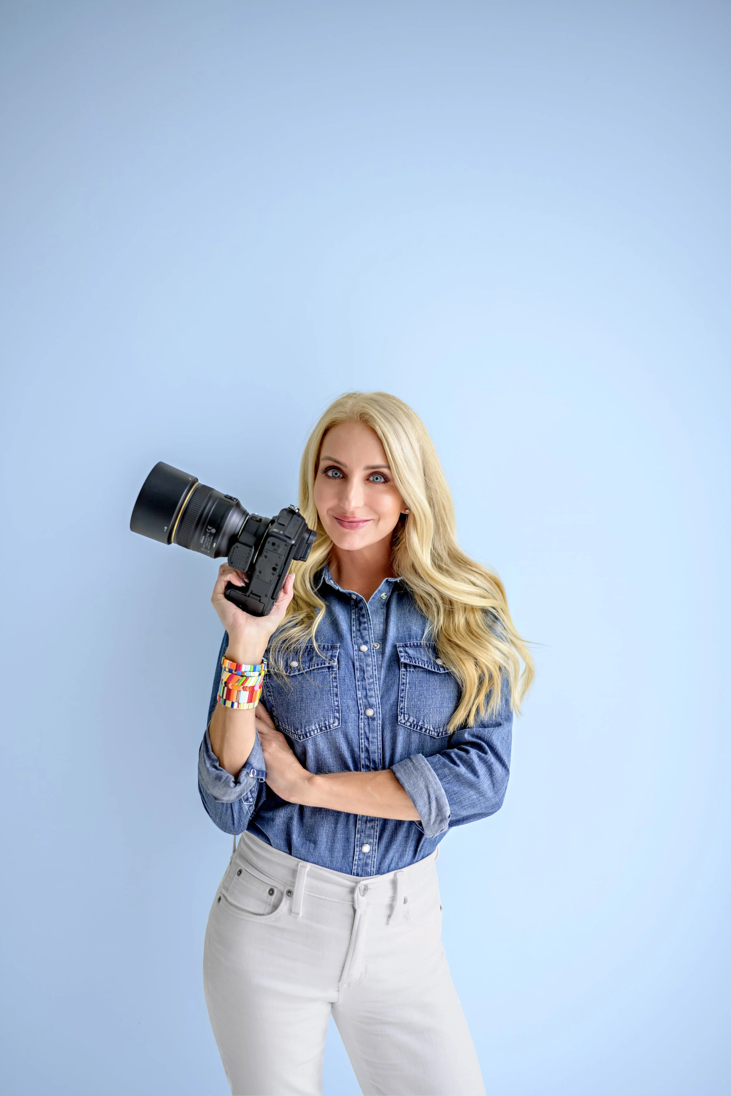 A woman with long blonde hair smiling while holding a camera with a large telephoto lens, standing against a light blue background, wearing a denim jacket, white pants, and colorful beaded bracelets.