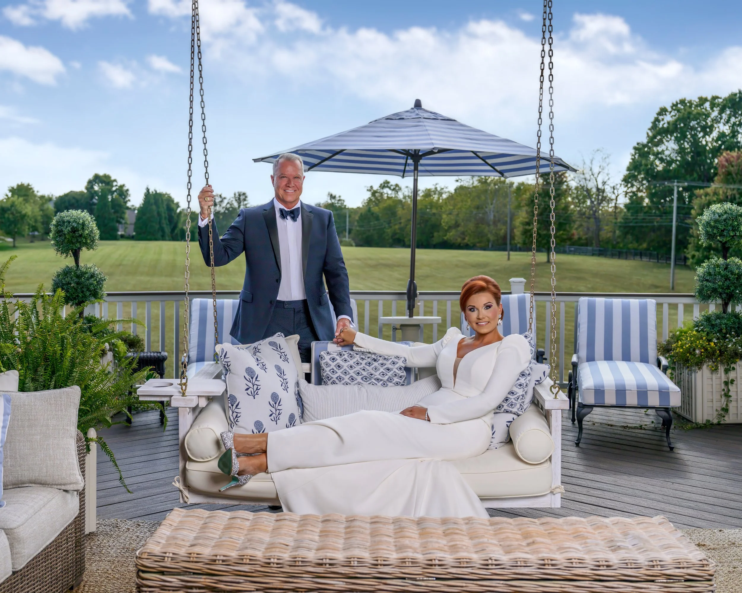 A man in a tuxedo and woman in a white dress sitting on a porch swing, shaking hands. The porch is decorated with potted plants and outdoor furniture. A blue and white striped umbrella provides shade, with a large green lawn and trees in the backgrou