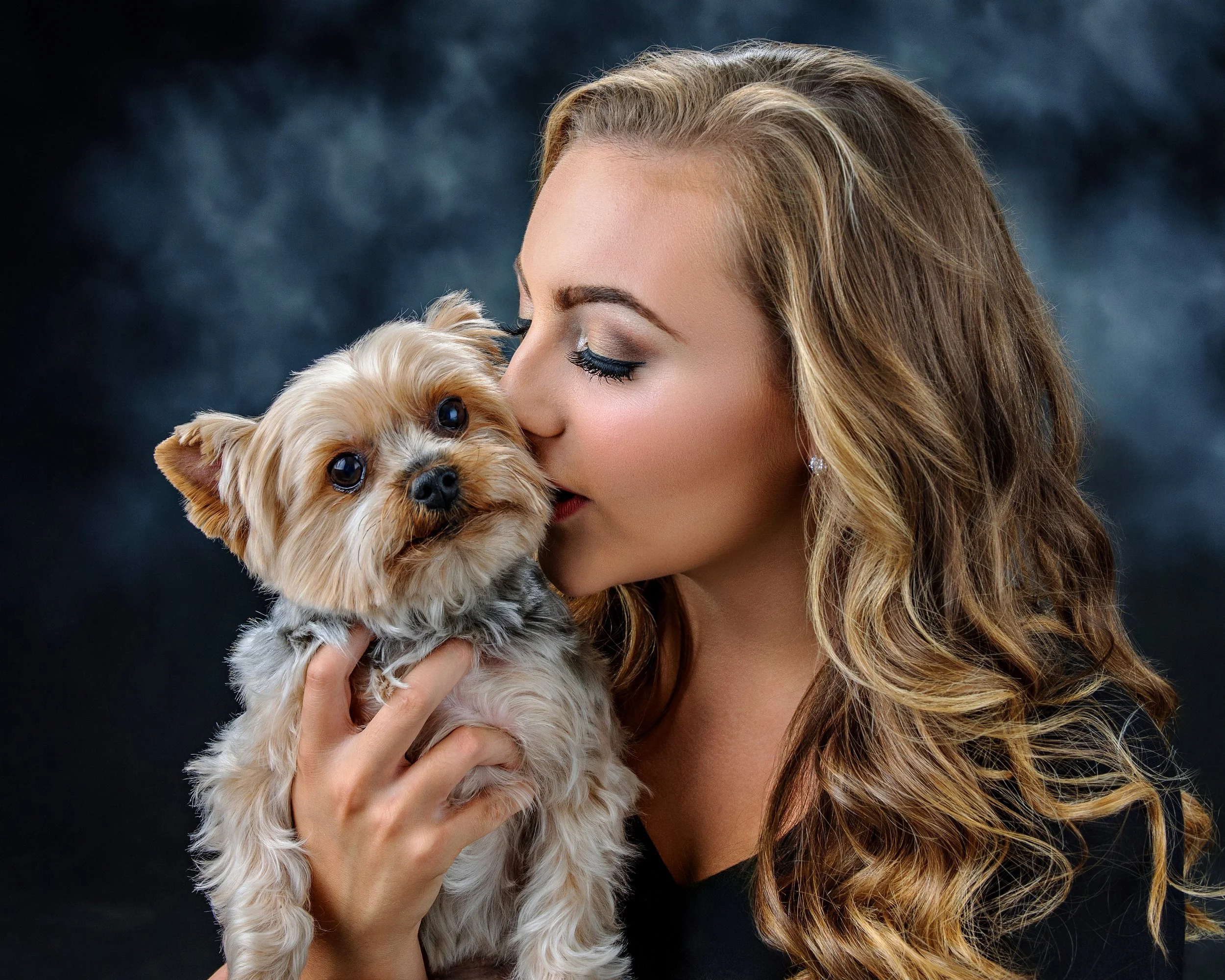 A woman with curly blonde hair holding a small dog with tan and gray fur, both close up against a dark, blurred background.